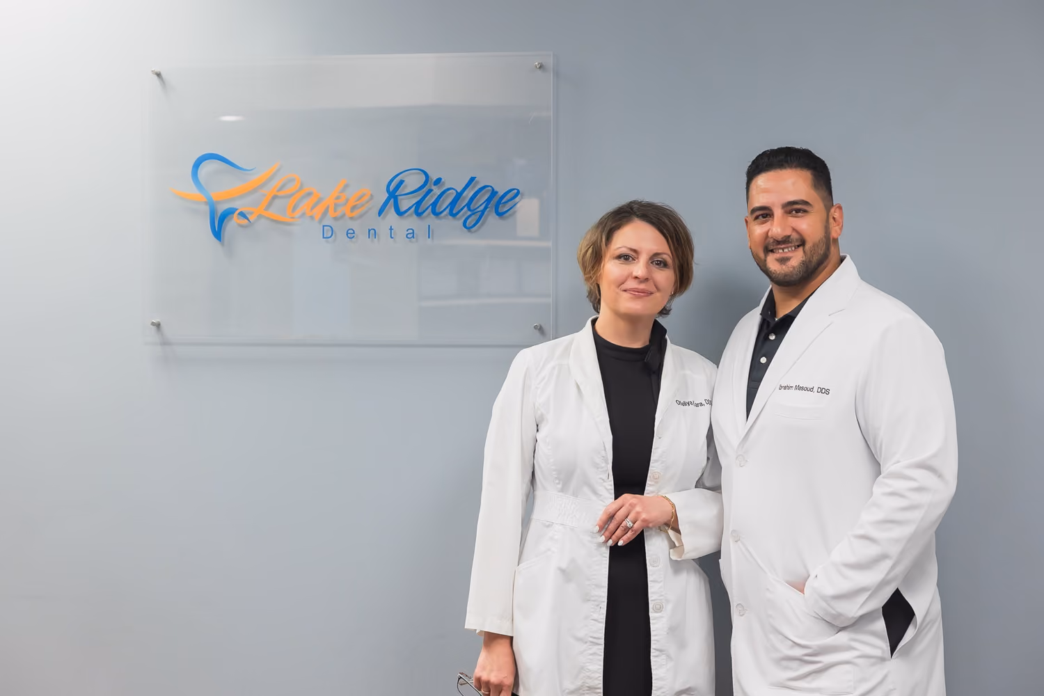 Two dental professionals in white coats standing and smiling in front of a Lake Ridge Dental sign on a gray wall.