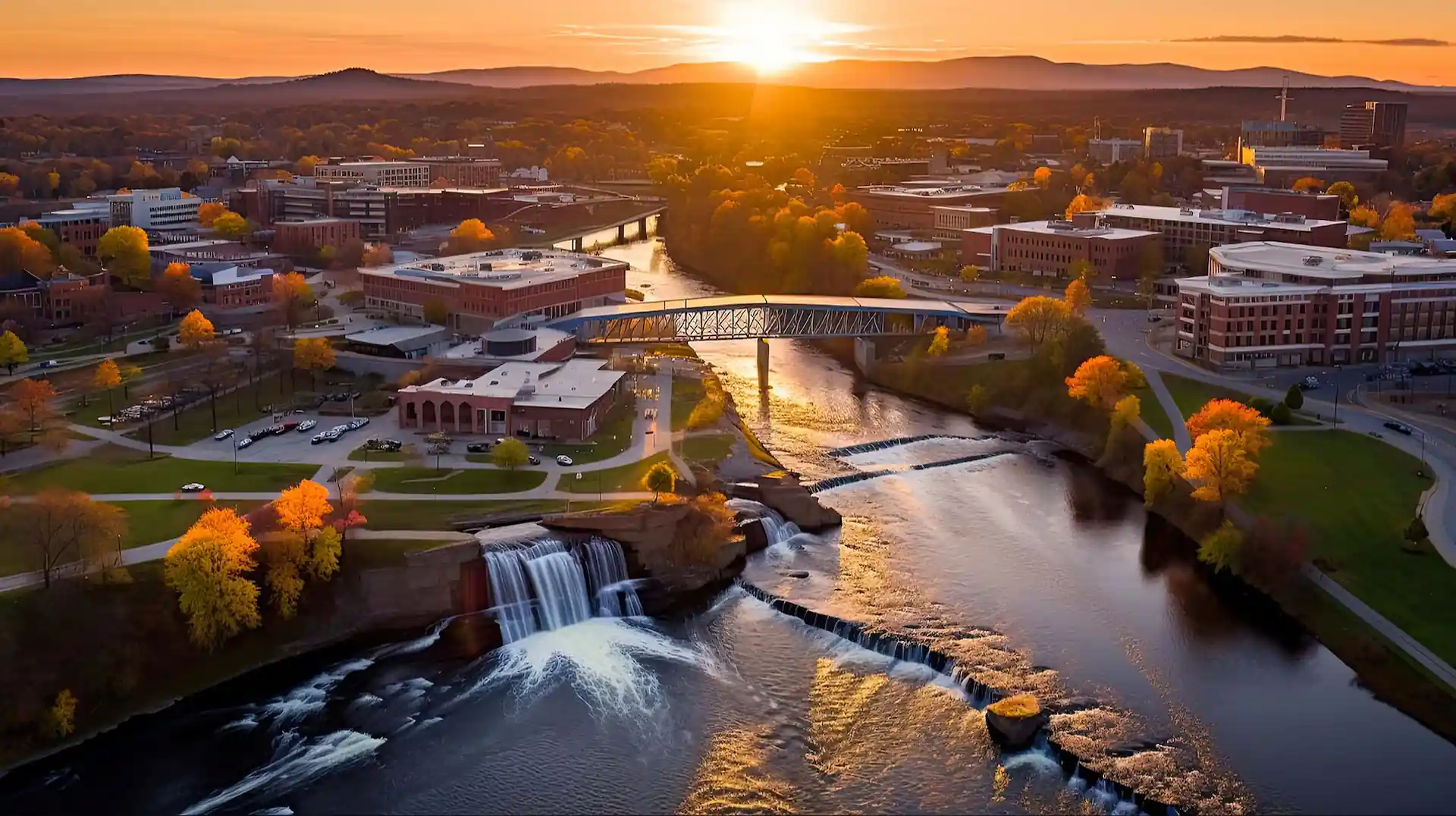 Sunset over a river with small waterfalls, a bridge, autumn trees, and buildings along the riverbank.