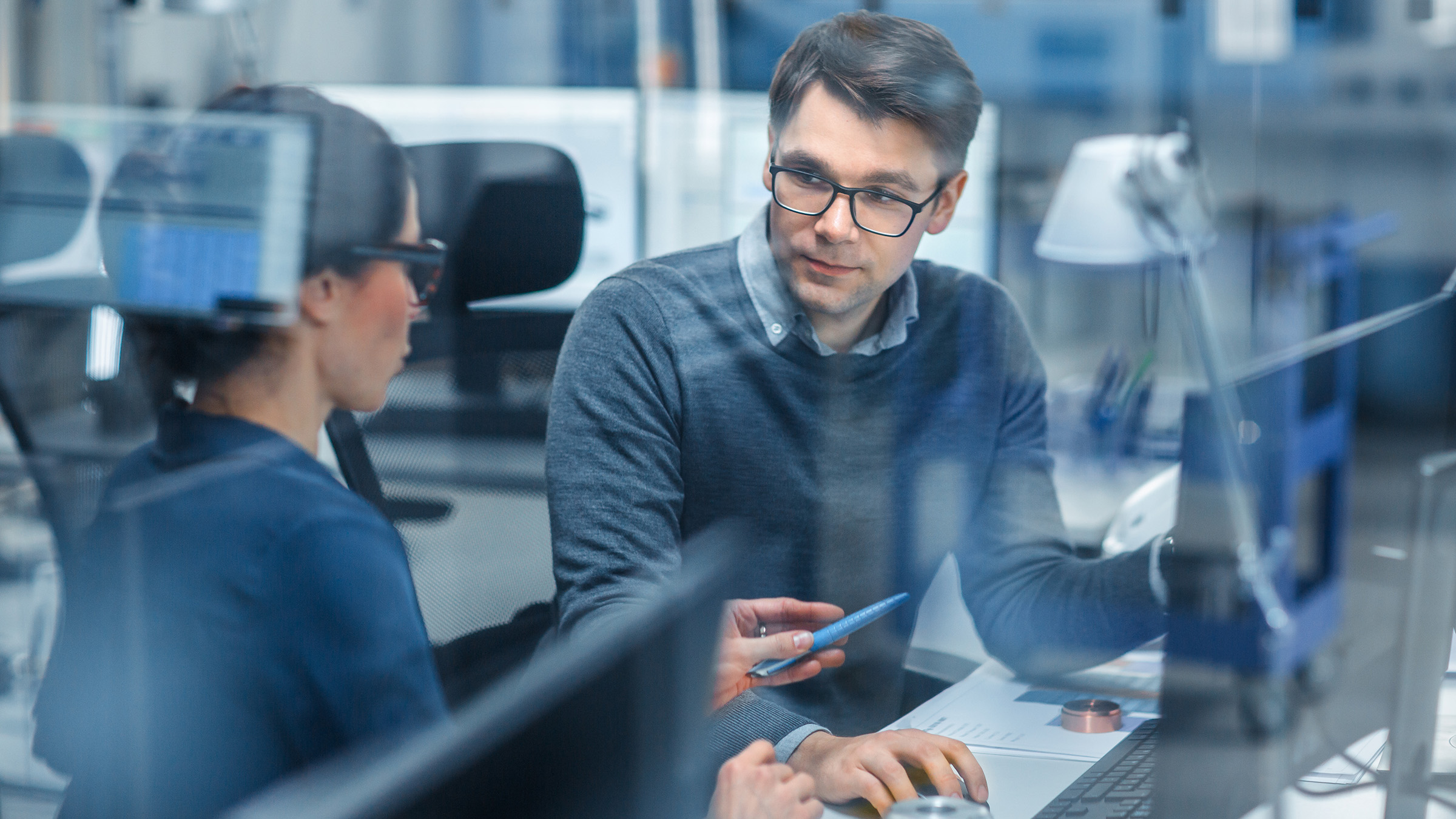Two professionals in a modern office setting having a focused discussion while working on a computer, viewed through a glass partition.