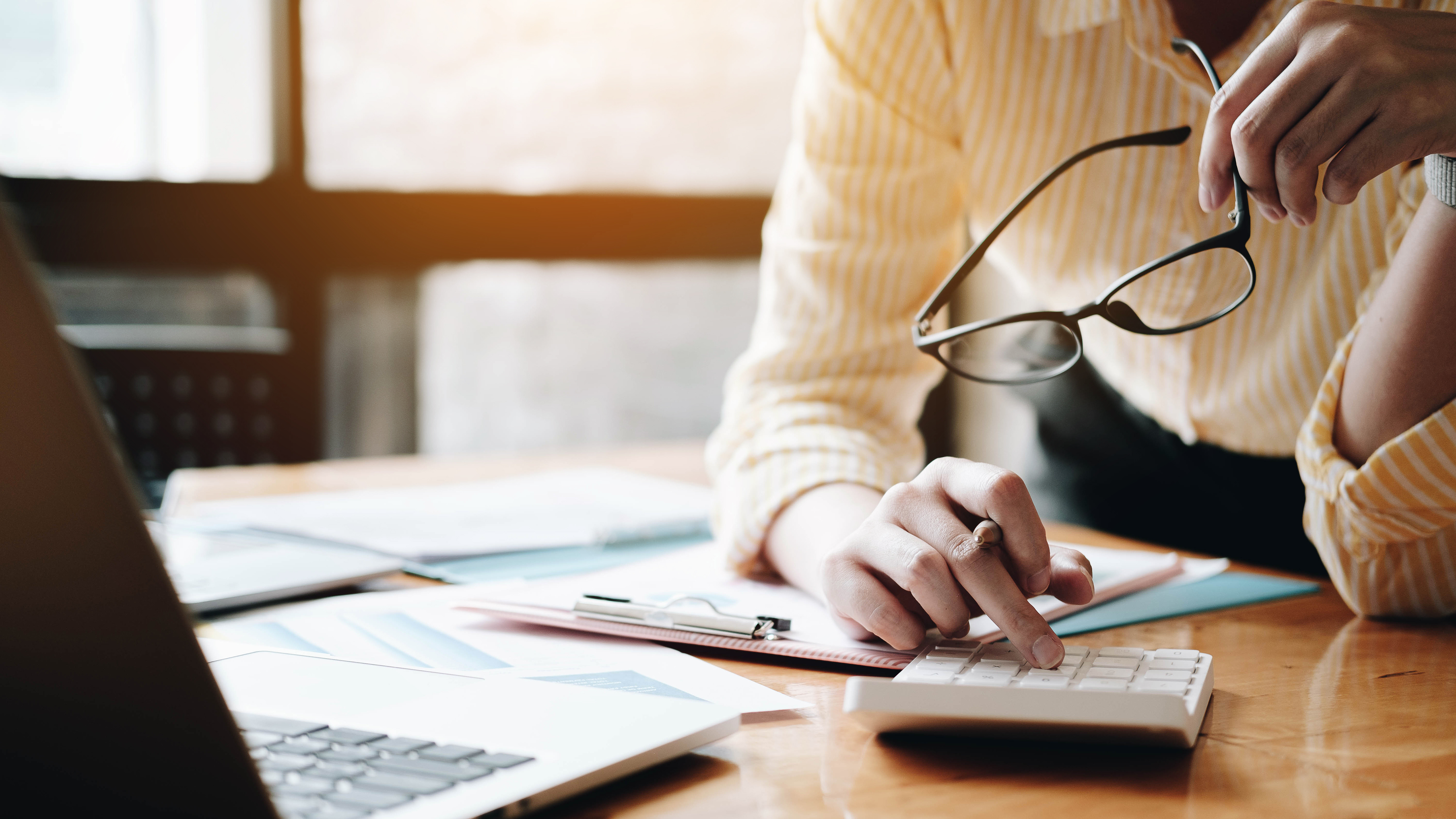 Close-up of a person using a calculator at a desk with documents, a laptop, and holding eyeglasses, representing financial or accounting work.