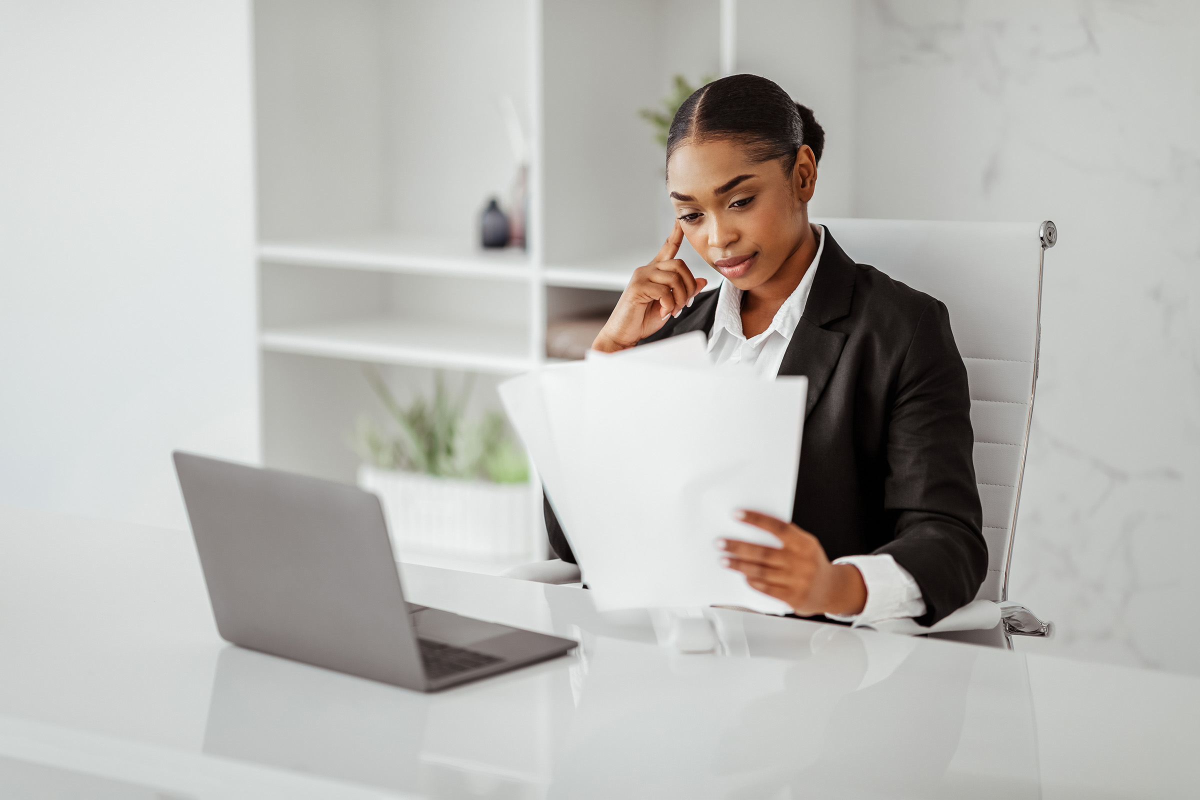 Focused businesswoman in a modern office reviewing documents while sitting at a desk with a laptop.