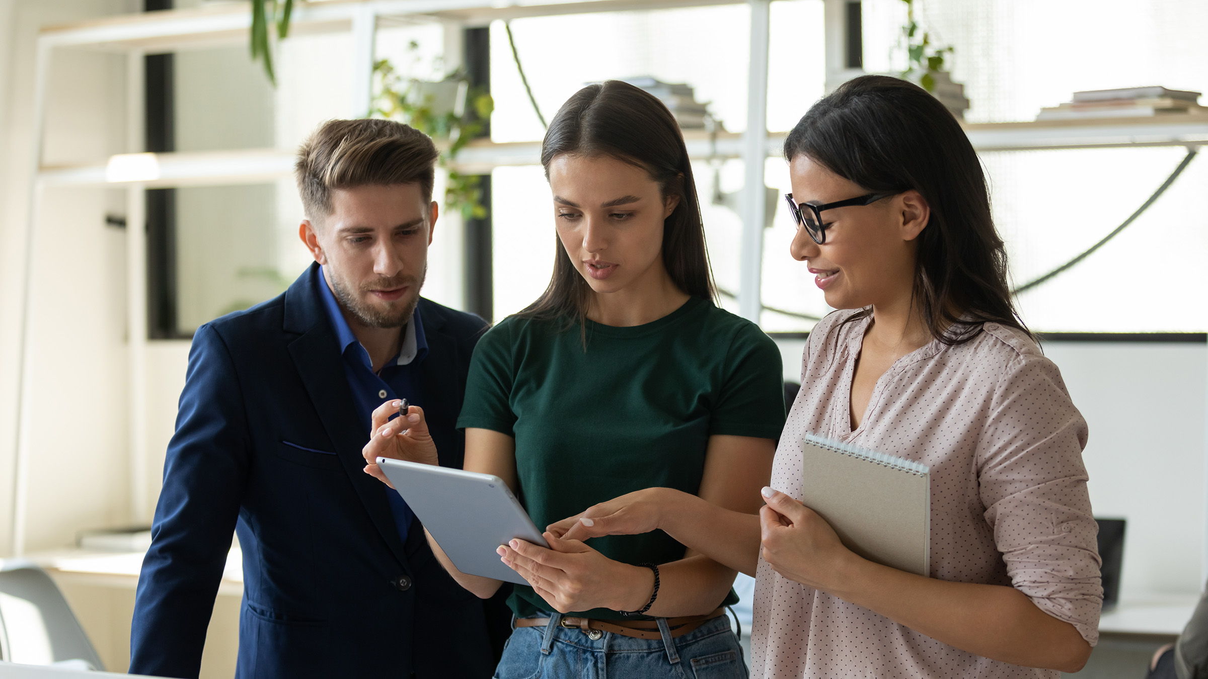 Three professionals collaborating in a modern office, reviewing information on a tablet and notebook, symbolizing teamwork and strategic planning.