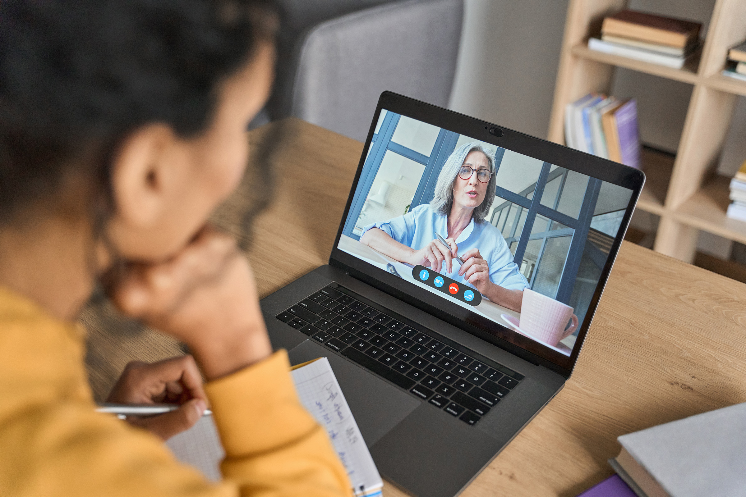 Woman taking notes while having a video call with a professional colleague on a laptop in a home office setting.