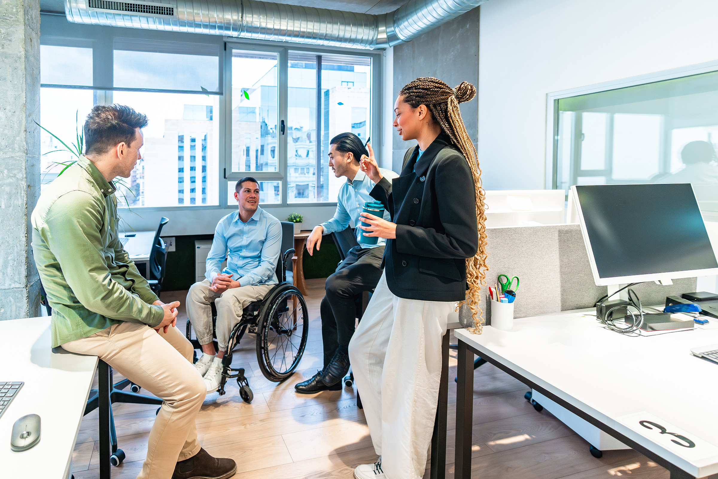 Diverse group of professionals, including a person in a wheelchair, having a casual conversation in a modern, inclusive office environment.