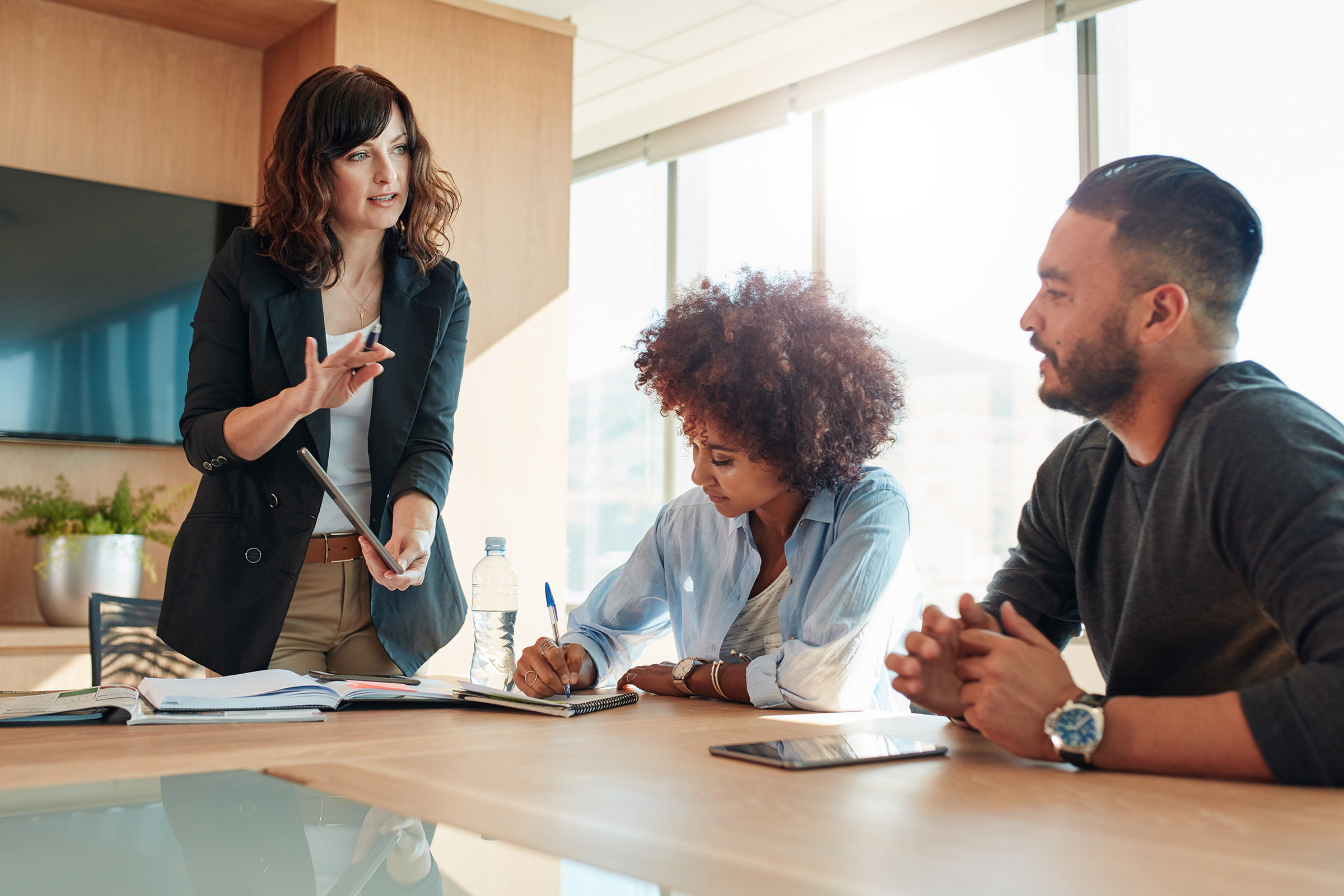 Businesswoman presenting ideas to colleagues during a team meeting in a modern office, with one team member taking notes and another listening attentively.