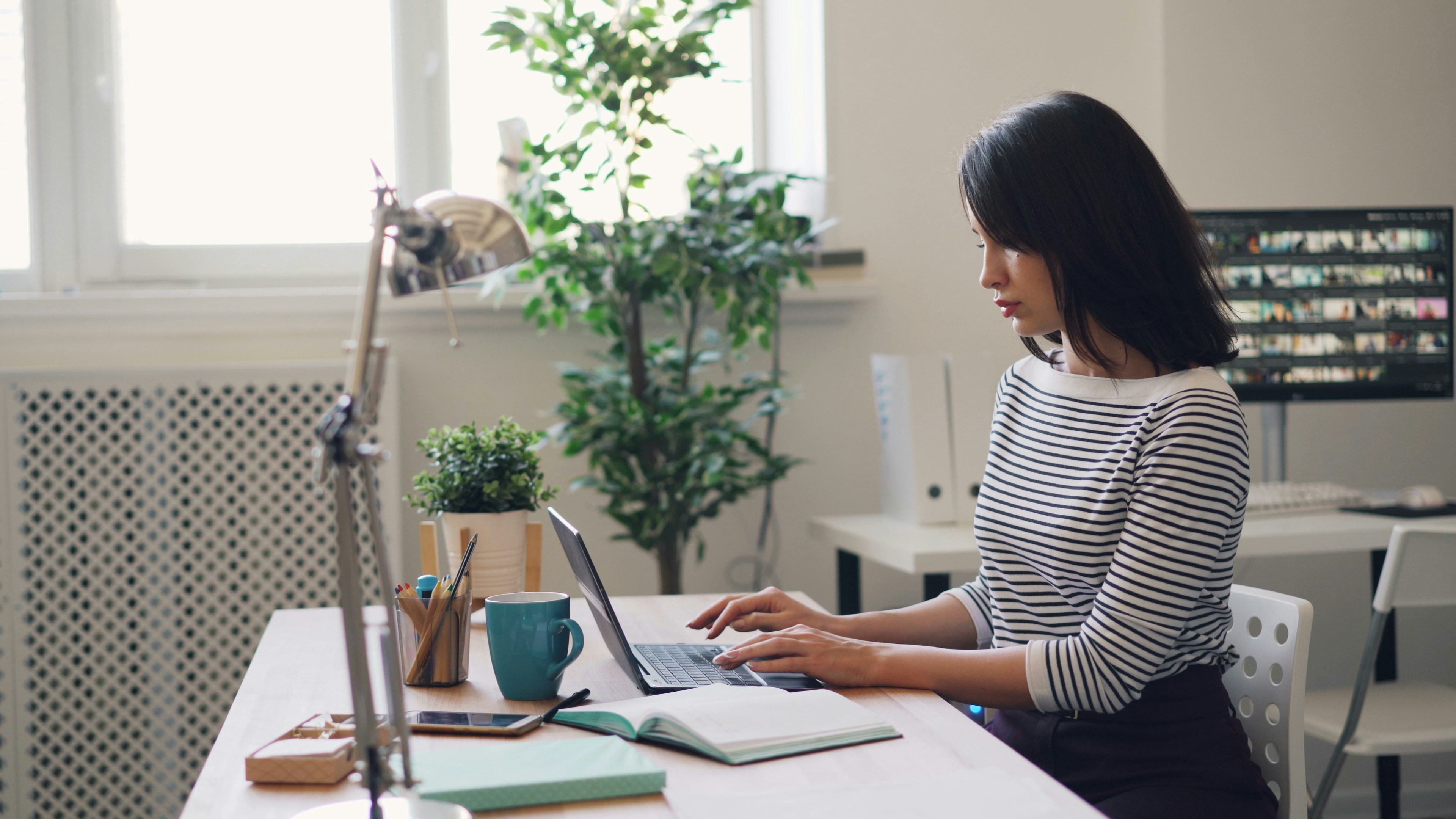 Young woman working on a laptop at a desk in a bright home office, surrounded by plants, notebooks, and office supplies.