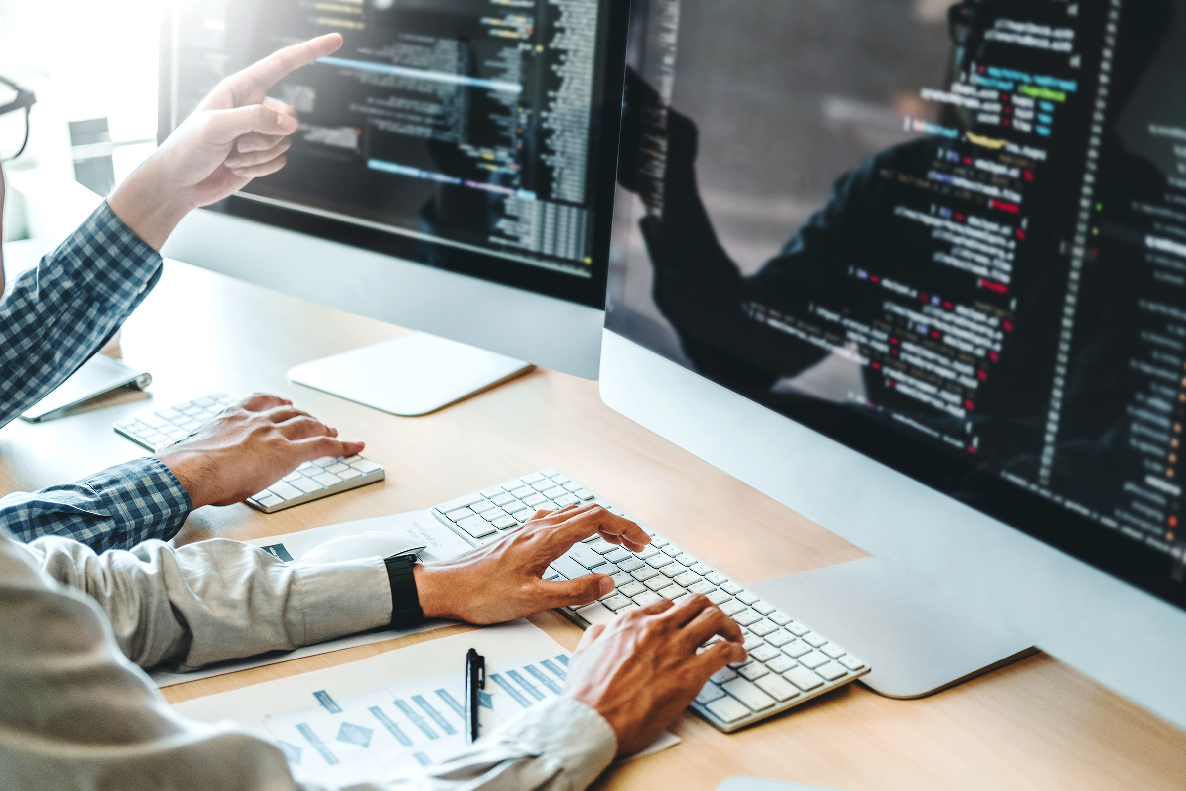 Two software engineers working together at a desk with dual monitors, reviewing and writing code in a collaborative programming environment.