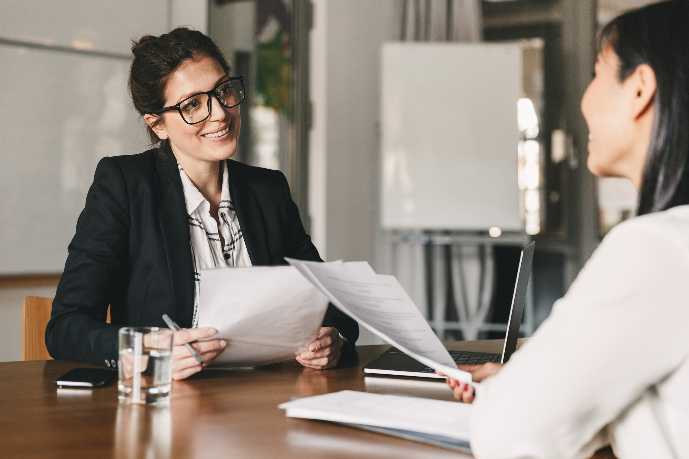 Smiling businesswoman in glasses conducting a job interview with a candidate across the table, reviewing a printed resume.