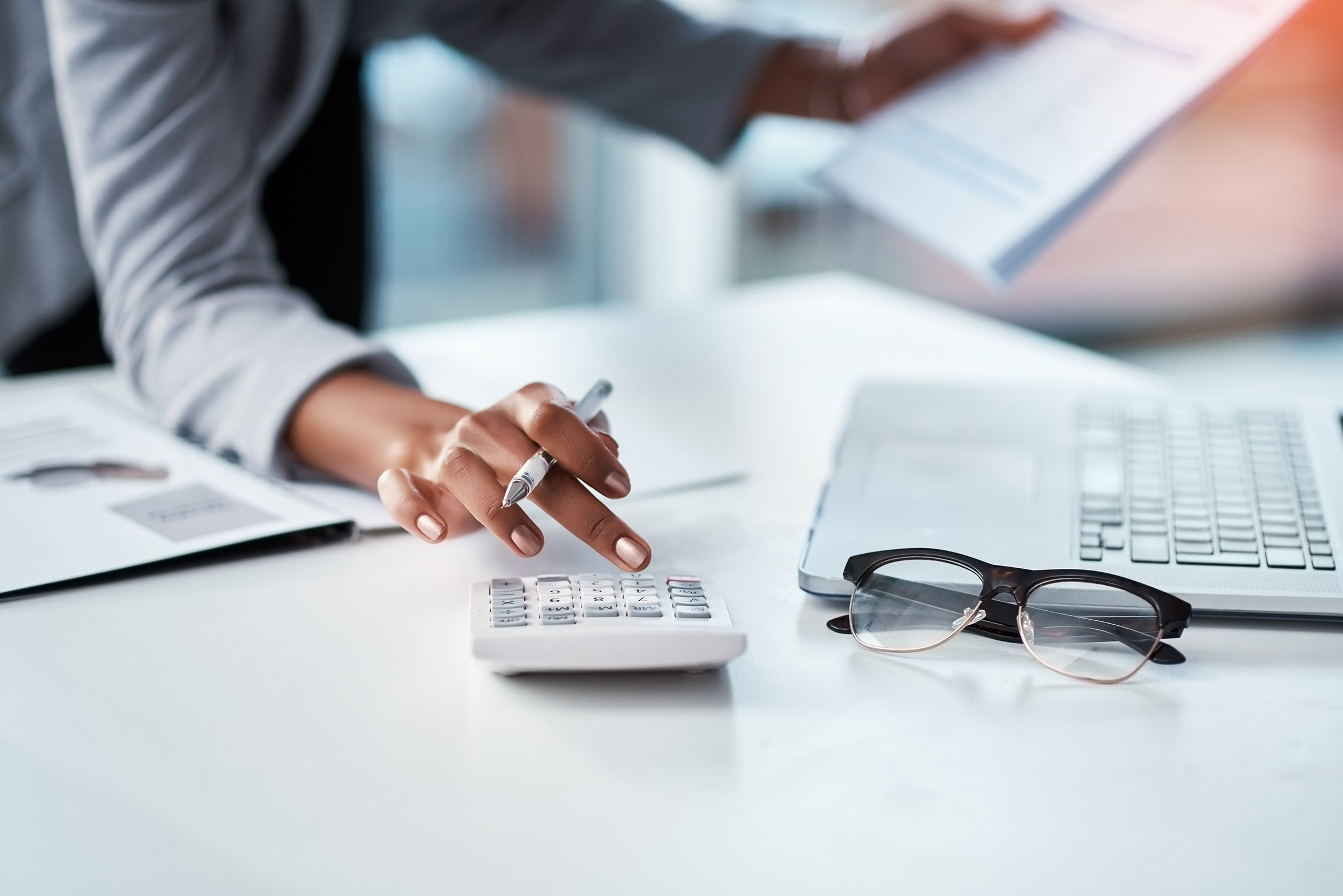 Close-up of a professional woman using a calculator at her desk, holding a document, with a laptop and eyeglasses nearby, representing financial or payroll work.