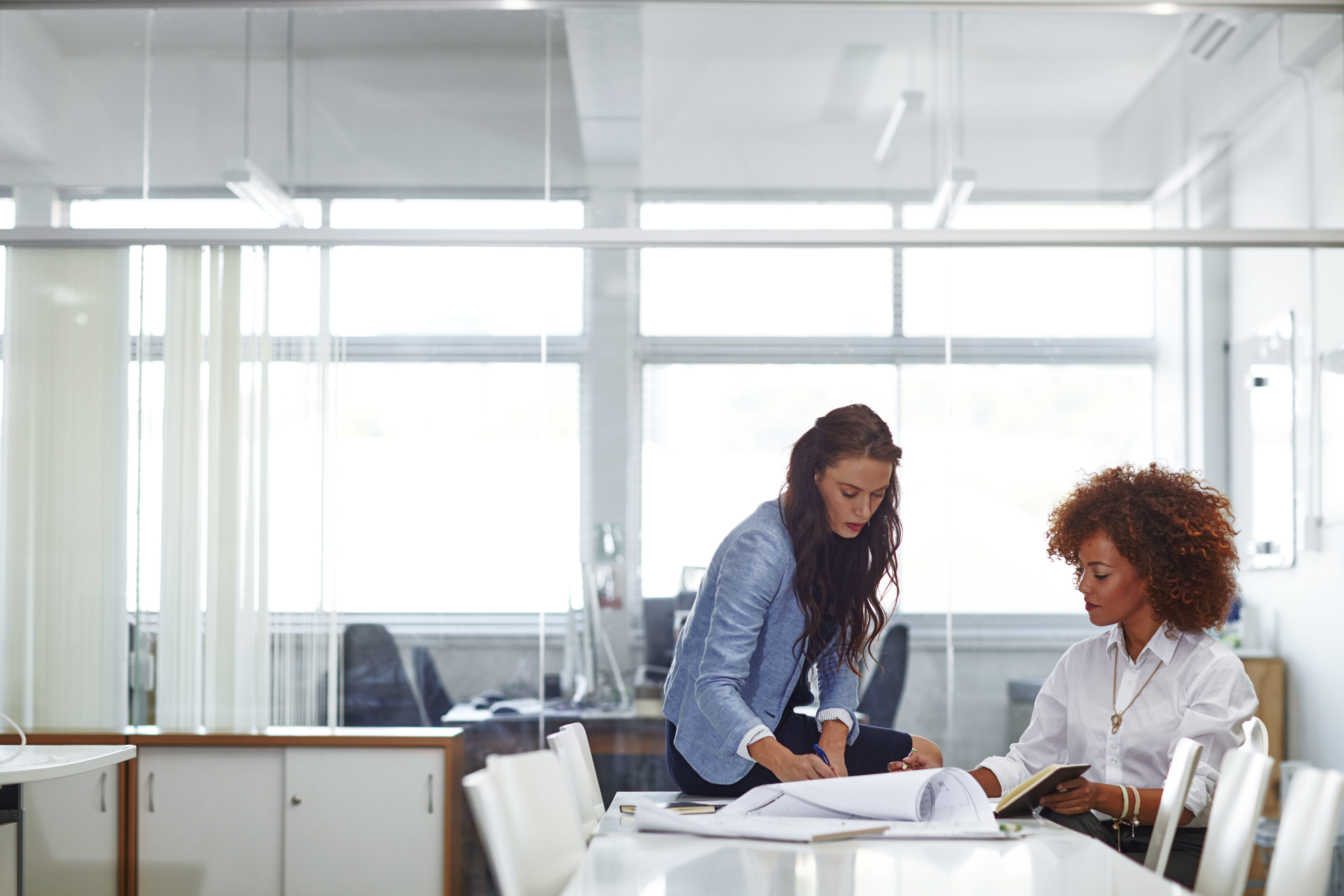 Two professional women collaborating in a modern office, reviewing architectural blueprints and notes together at a conference table.