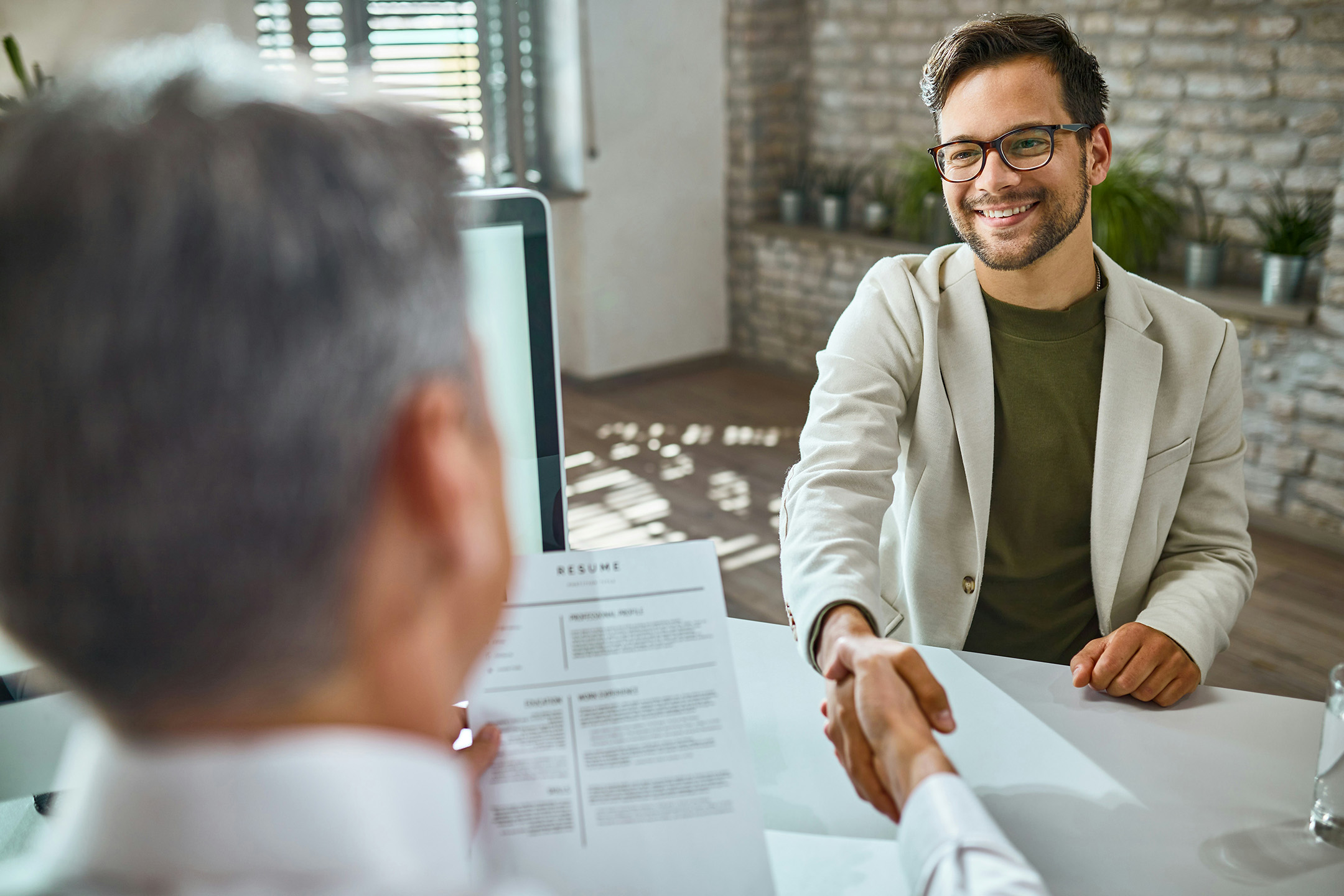 Smiling job candidate shaking hands with an interviewer during a successful interview, with a resume and computer visible on the desk.