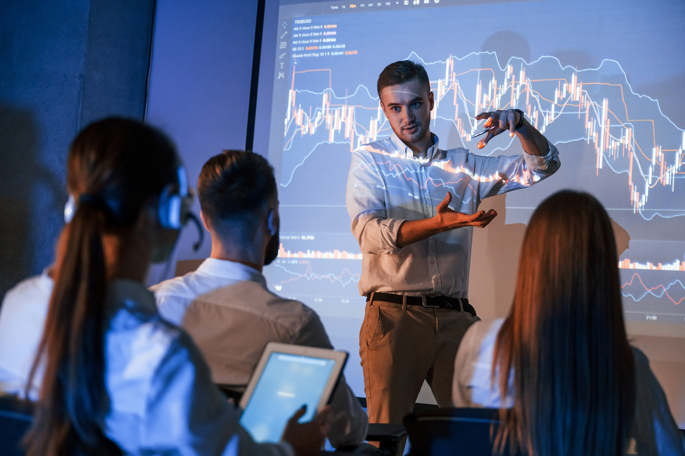 A presenter explains business charts projected on a screen to a small group of colleagues.