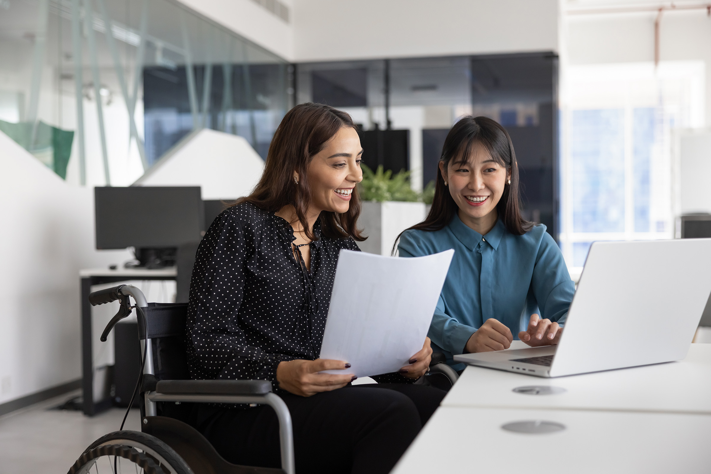 Two colleagues reviewing a document and working together on a laptop in a modern office, with one woman seated in a wheelchair.