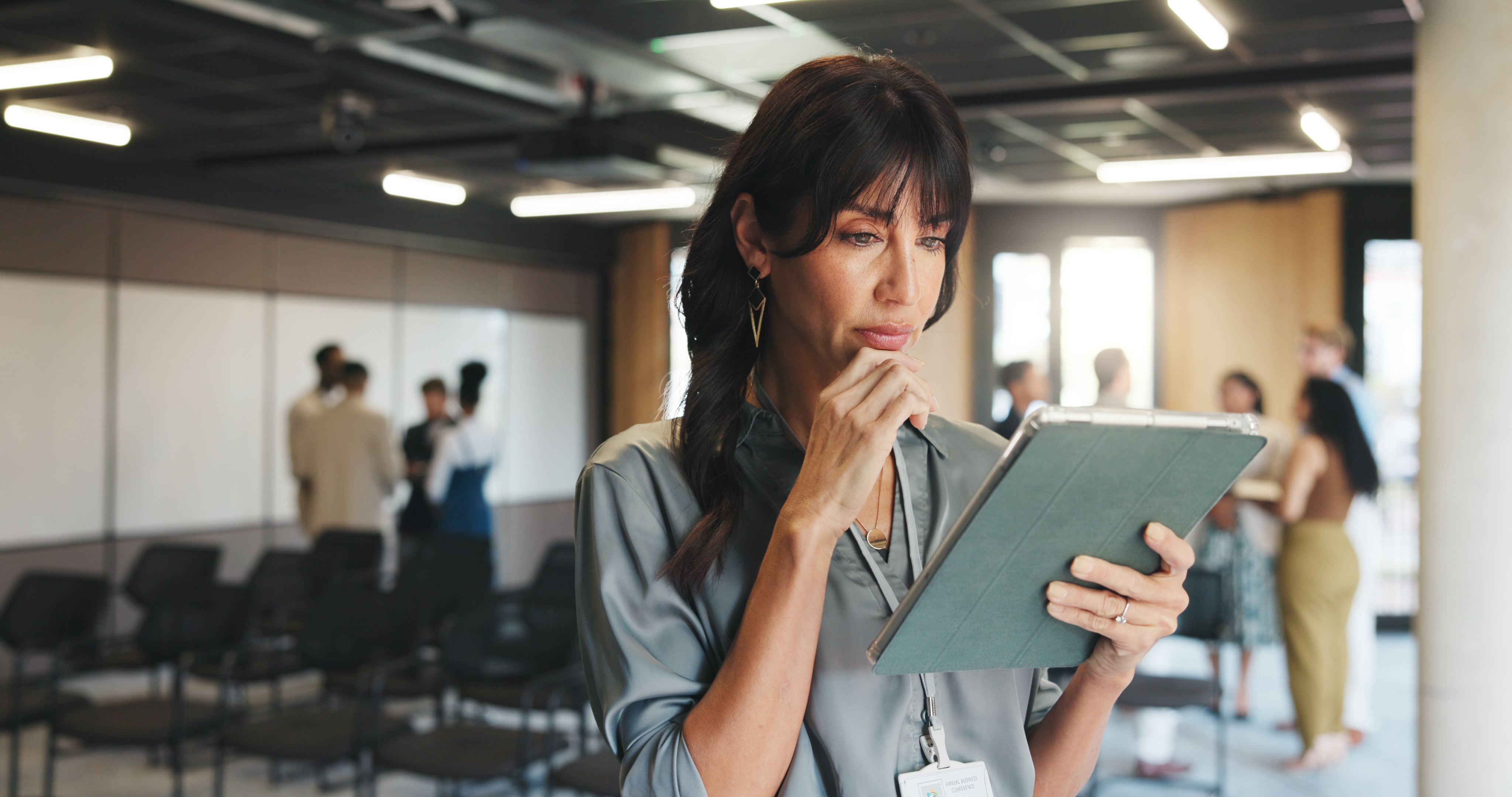 Professional reviewing information on a tablet in a conference room while colleagues collaborate in the background.