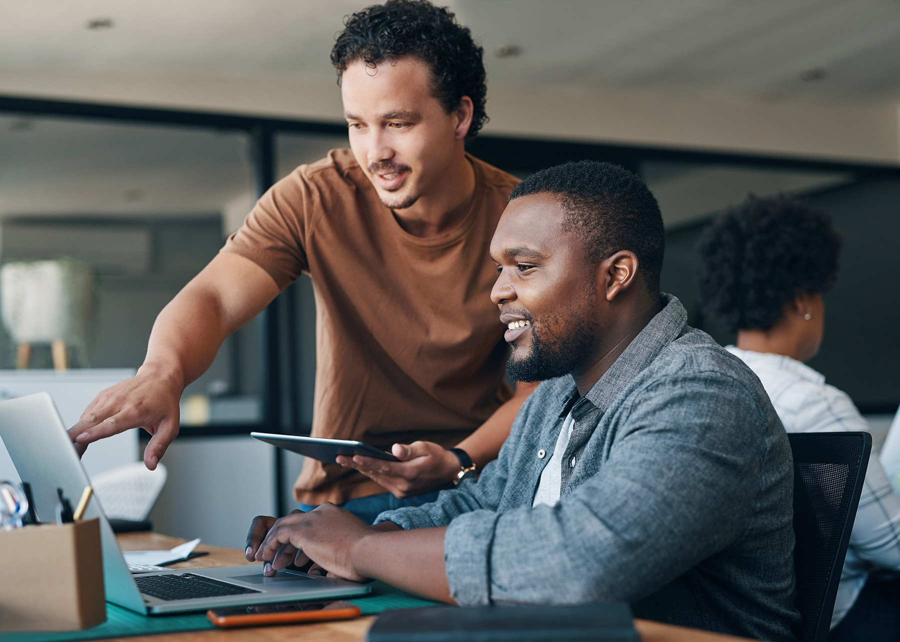 Two coworkers collaborating at a desk, reviewing work on a laptop and tablet in a modern office.