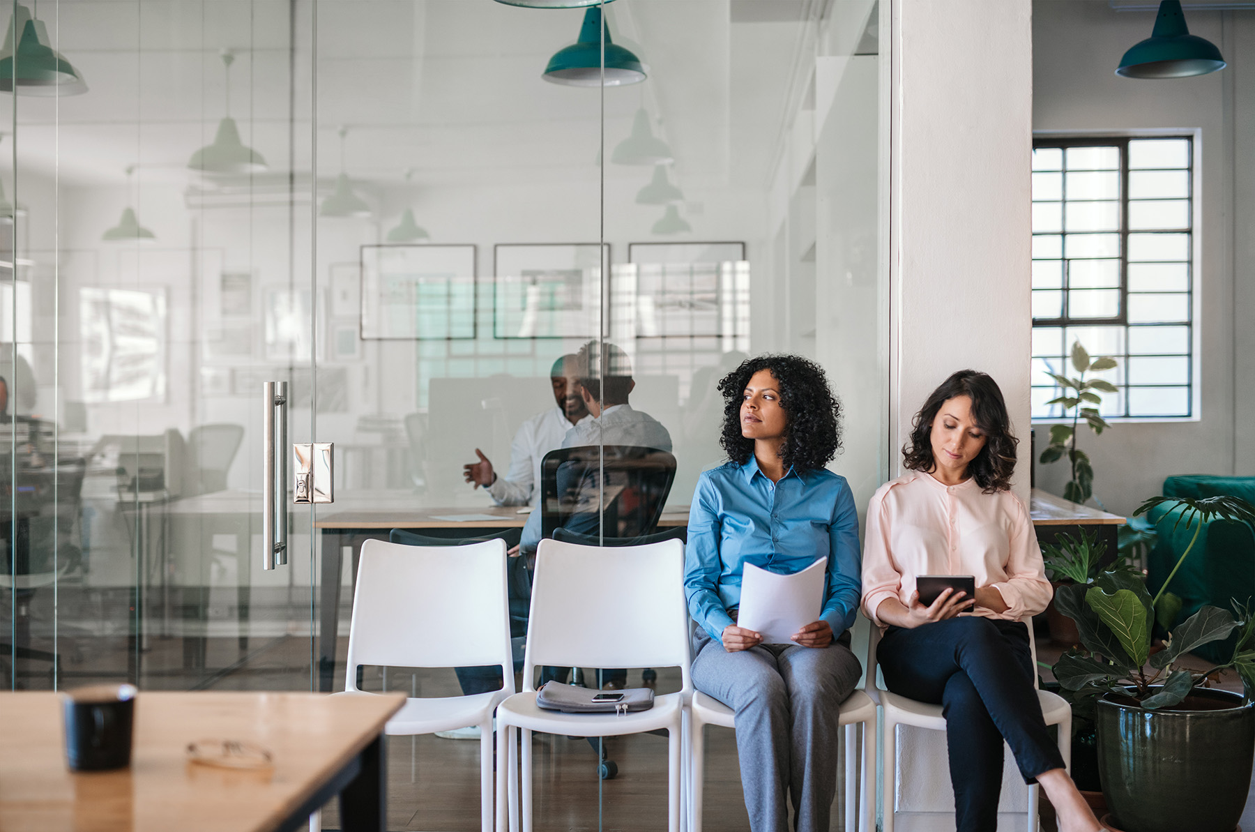 Two candidates sit in a modern office waiting area holding resumes, while an interview takes place behind a glass conference room.