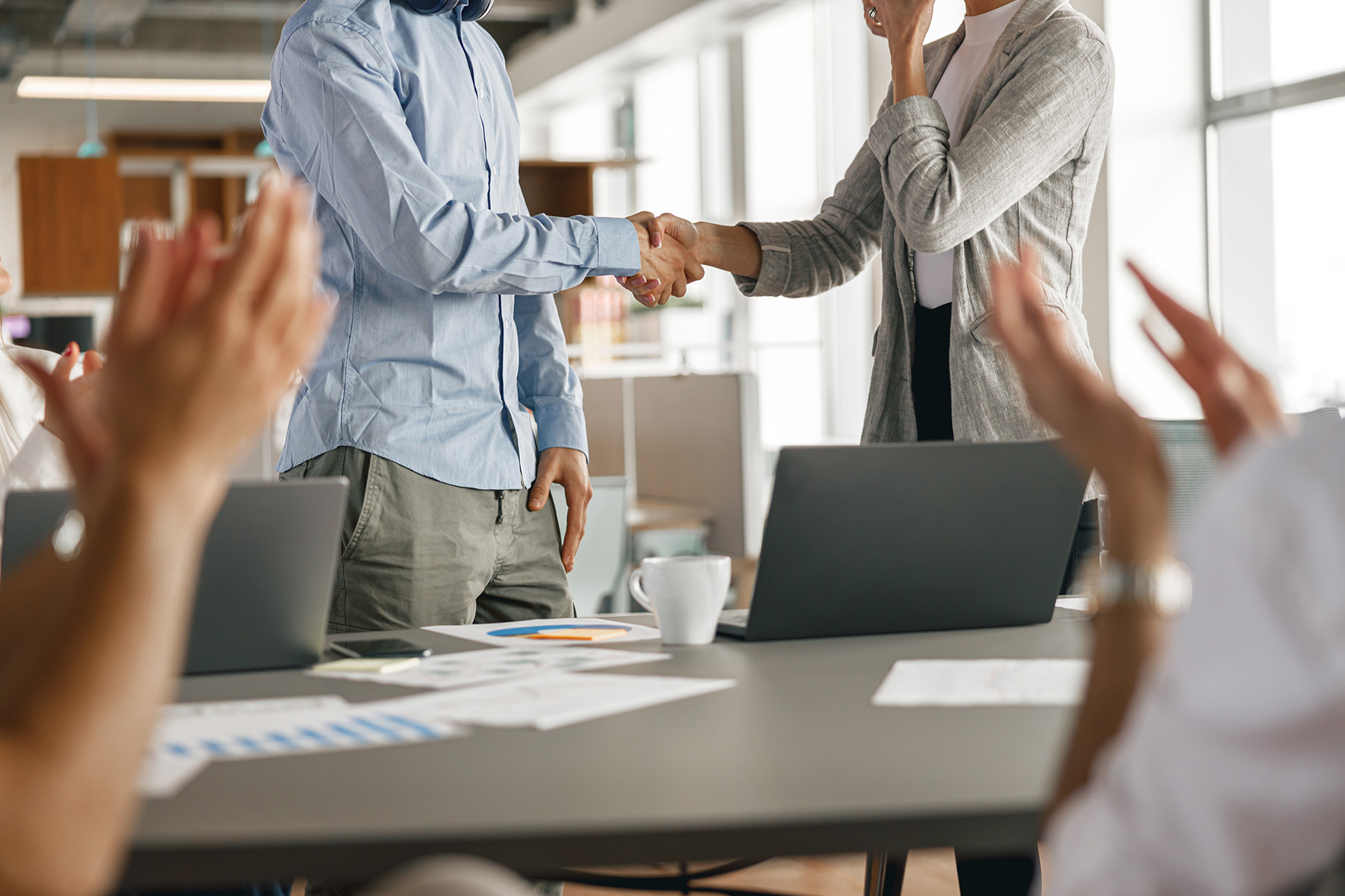 Two professionals shake hands in an office meeting room while colleagues applaud, signaling a successful hire or business agreement.