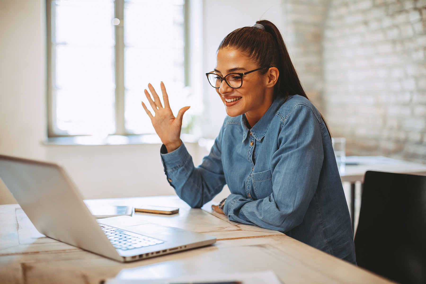 Professional woman smiling and waving during a video interview on a laptop in a bright home office setting.