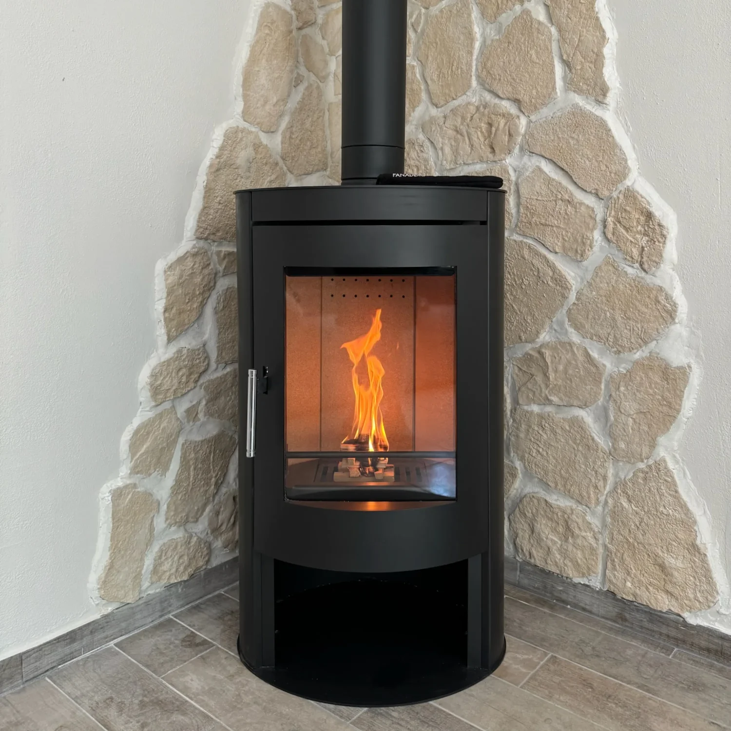 Poêle à bois noir avec une flamme visible, installé dans un coin avec mur en pierres.