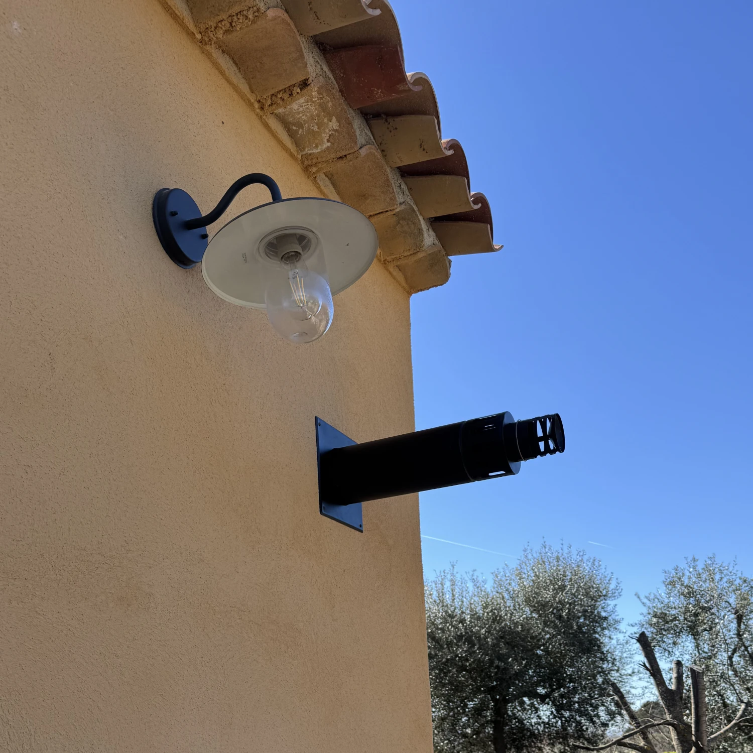 Mur beige avec une lampe extérieure et une cheminée noire contre un ciel bleu clair.