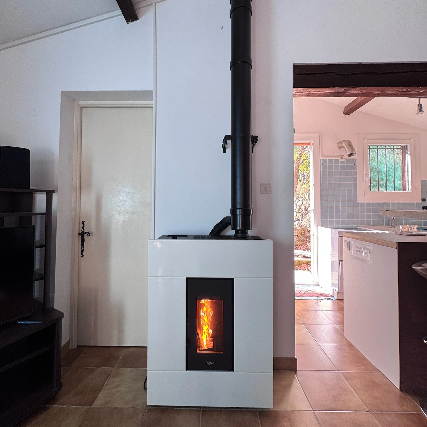 Poêle à granulés blanc allumé avec cheminée noire dans une pièce avec carrelage marron et cuisine adjacente.