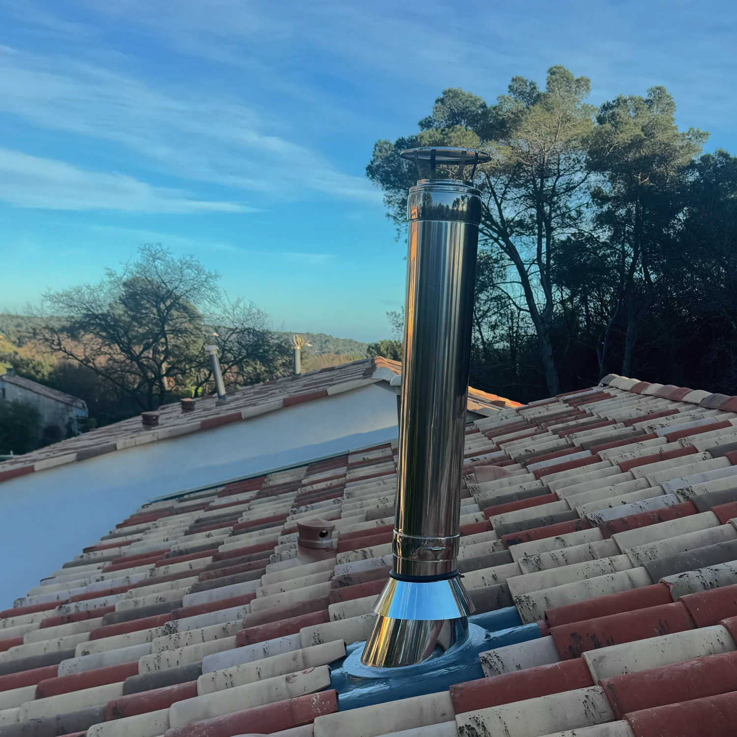 Conduit de cheminée en acier inoxydable sur un toit de tuiles rouges et beige sous un ciel bleu.
