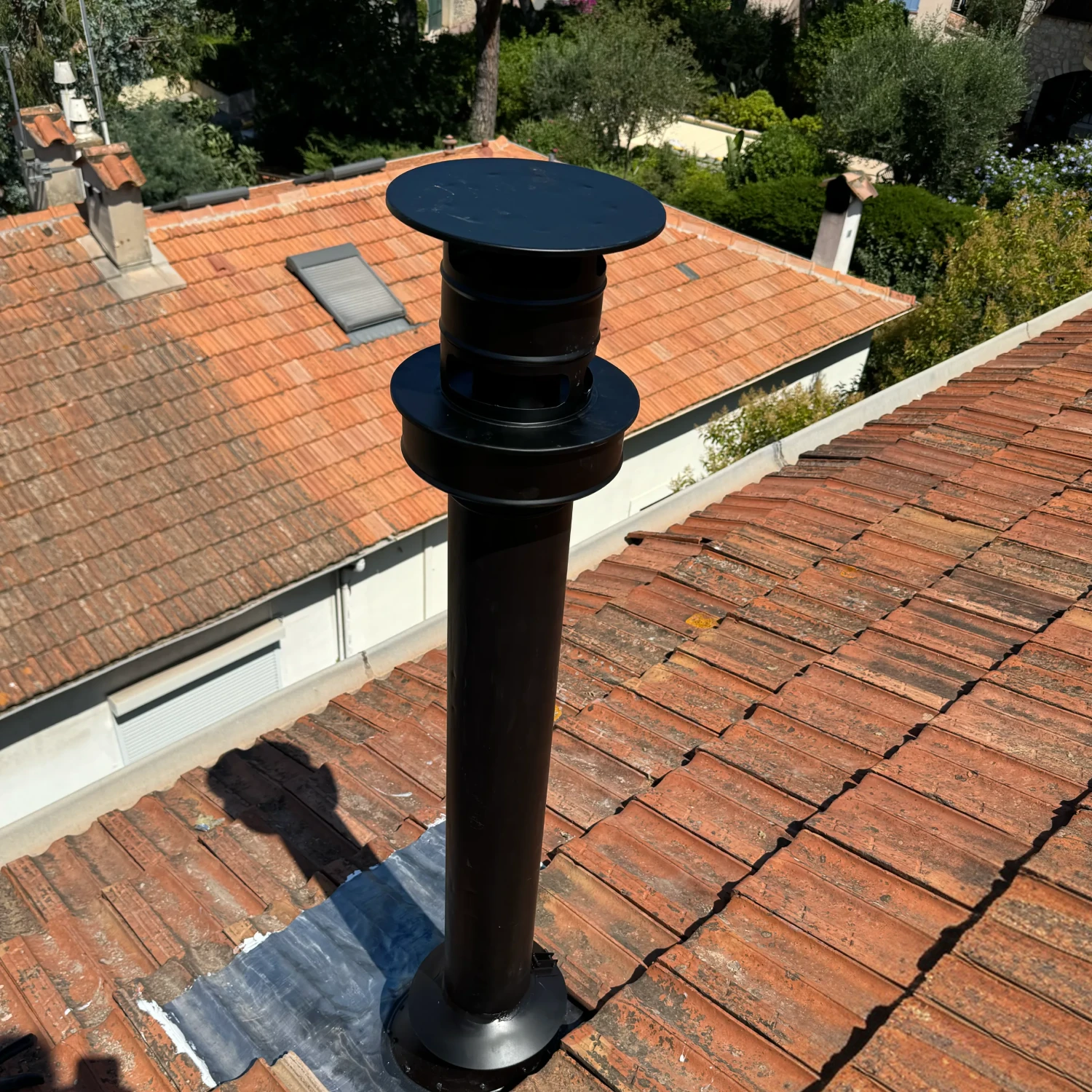Conduit d'aération noir double flux installé sur un toit en tuiles rouges sous un ciel clair.