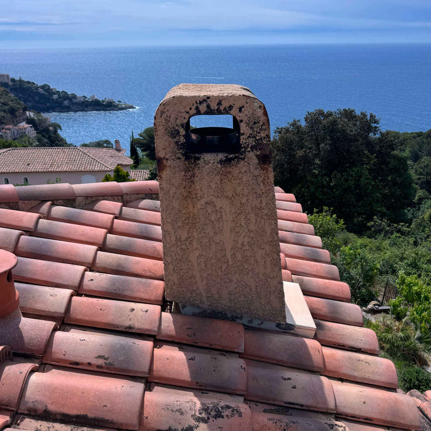 Cheminée en pierre sur un toit en tuiles rouges avec vue sur la mer et des arbres verts.