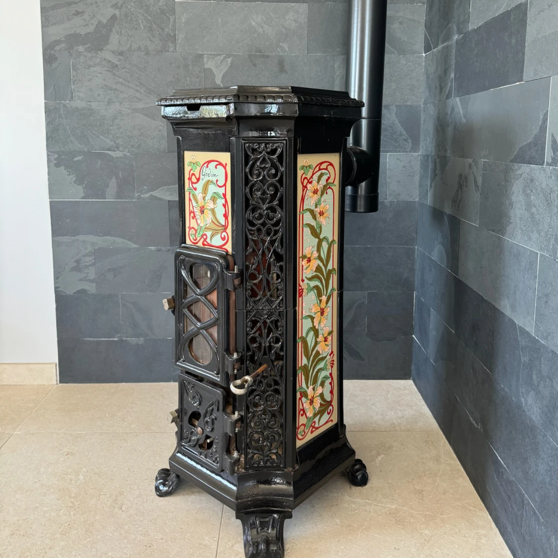 Poêle à bois ancien avec panneaux décoratifs floraux et porte en fonte noire, installé dans un coin au mur gris.