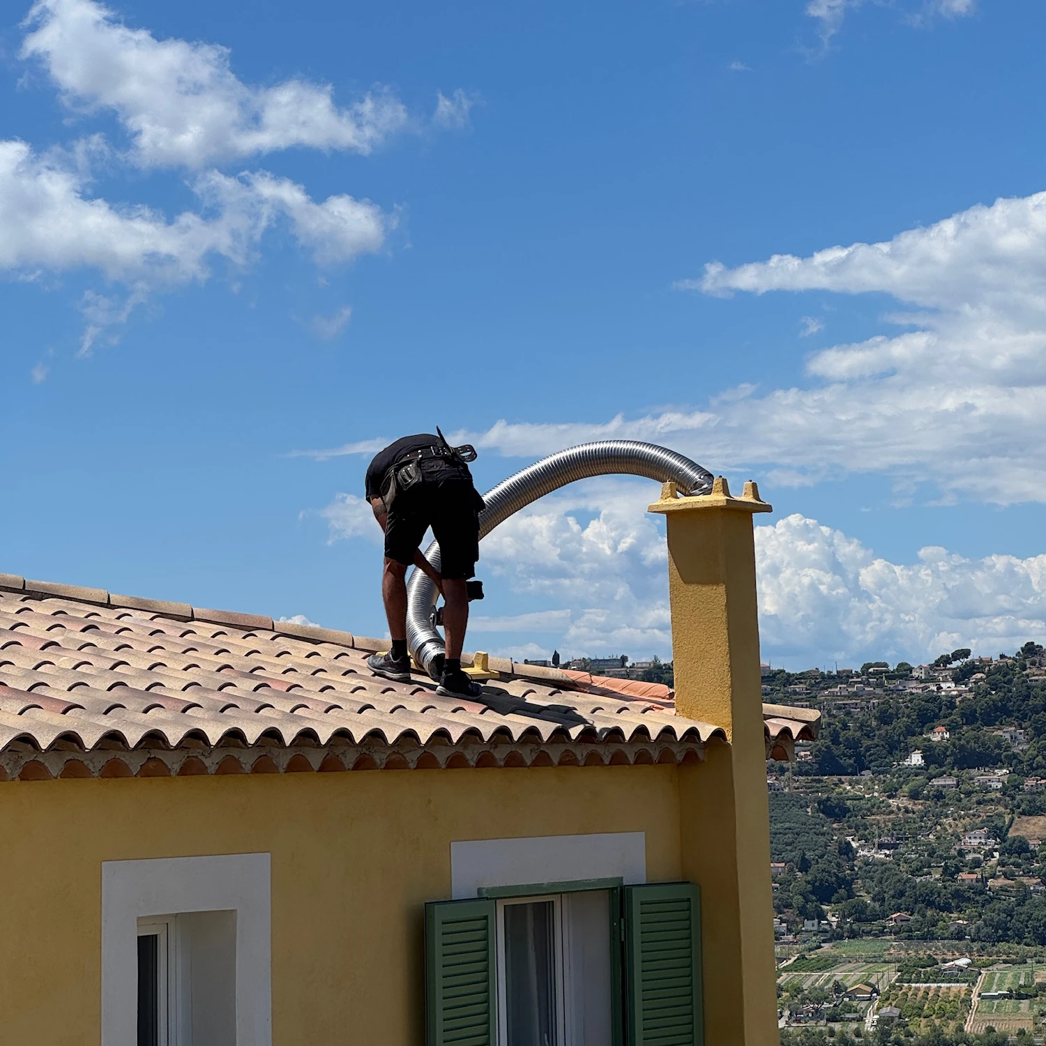 Un ouvrier remplace un conduit flexible sur le toit d'une maison jaune sous un ciel bleu.