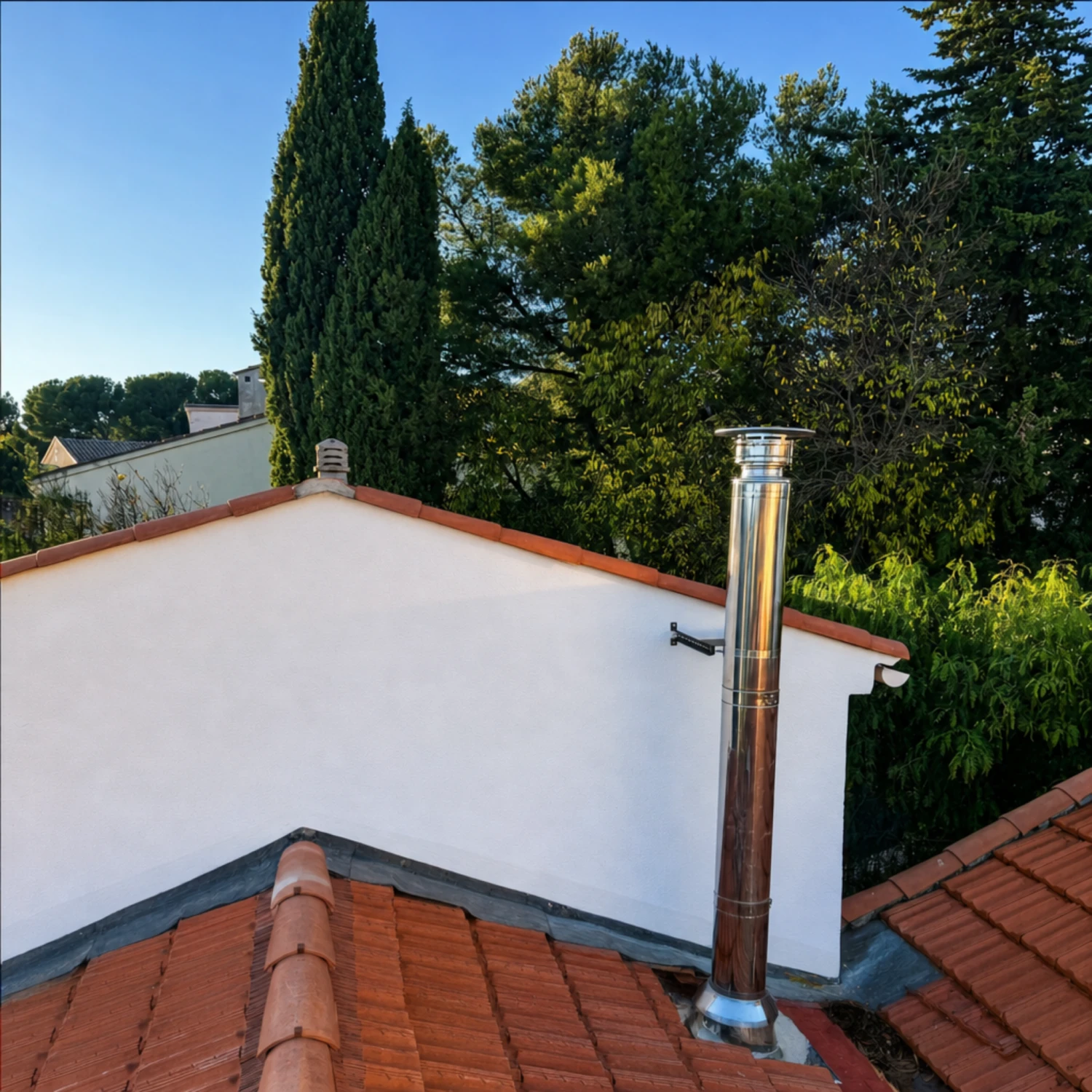 Cheminée inox brillante installée sur un toit en tuiles rouges avec des arbres verts autour.