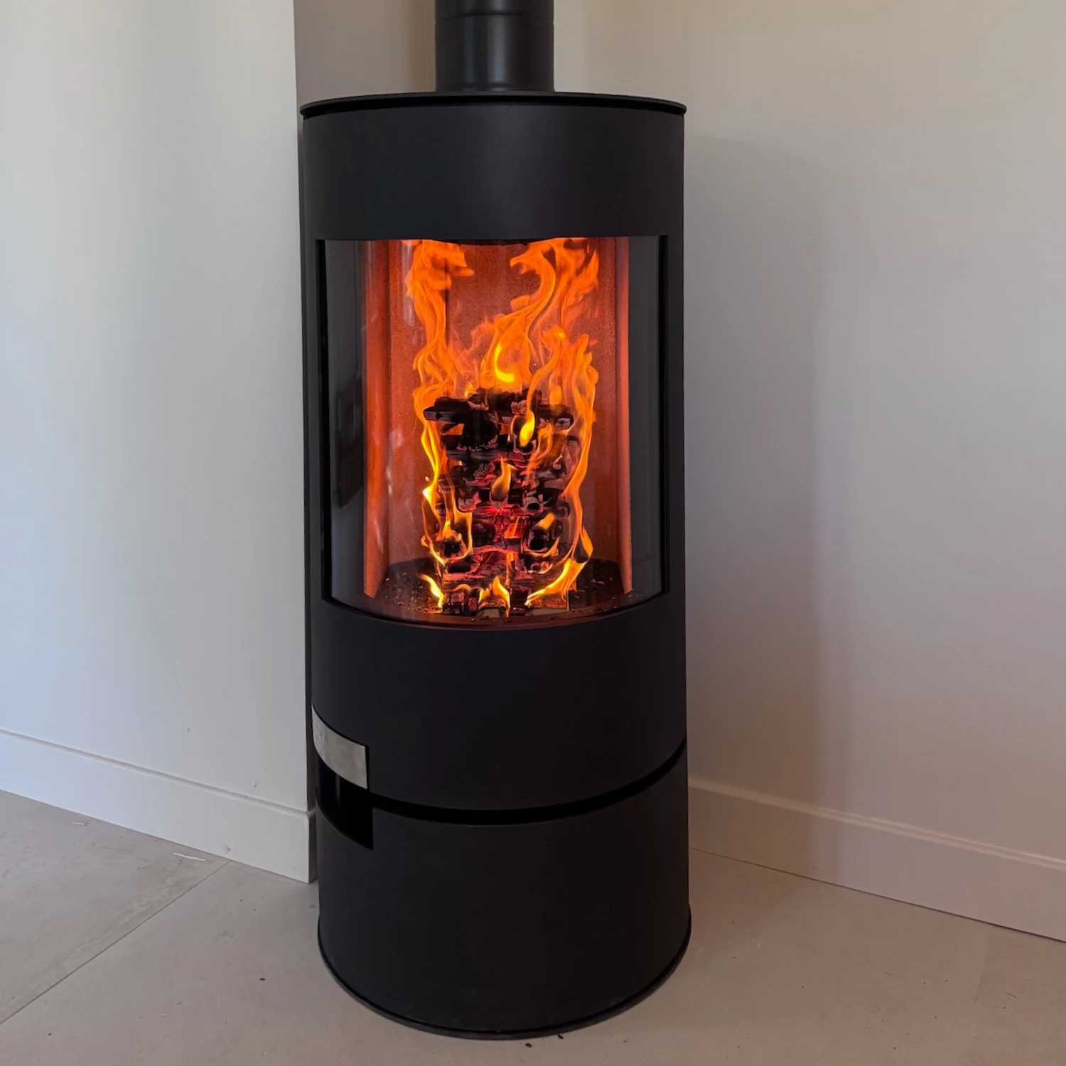 Poêle à bois noir cylindrique avec feu visible derrière une vitre, placé dans un coin de mur blanc.