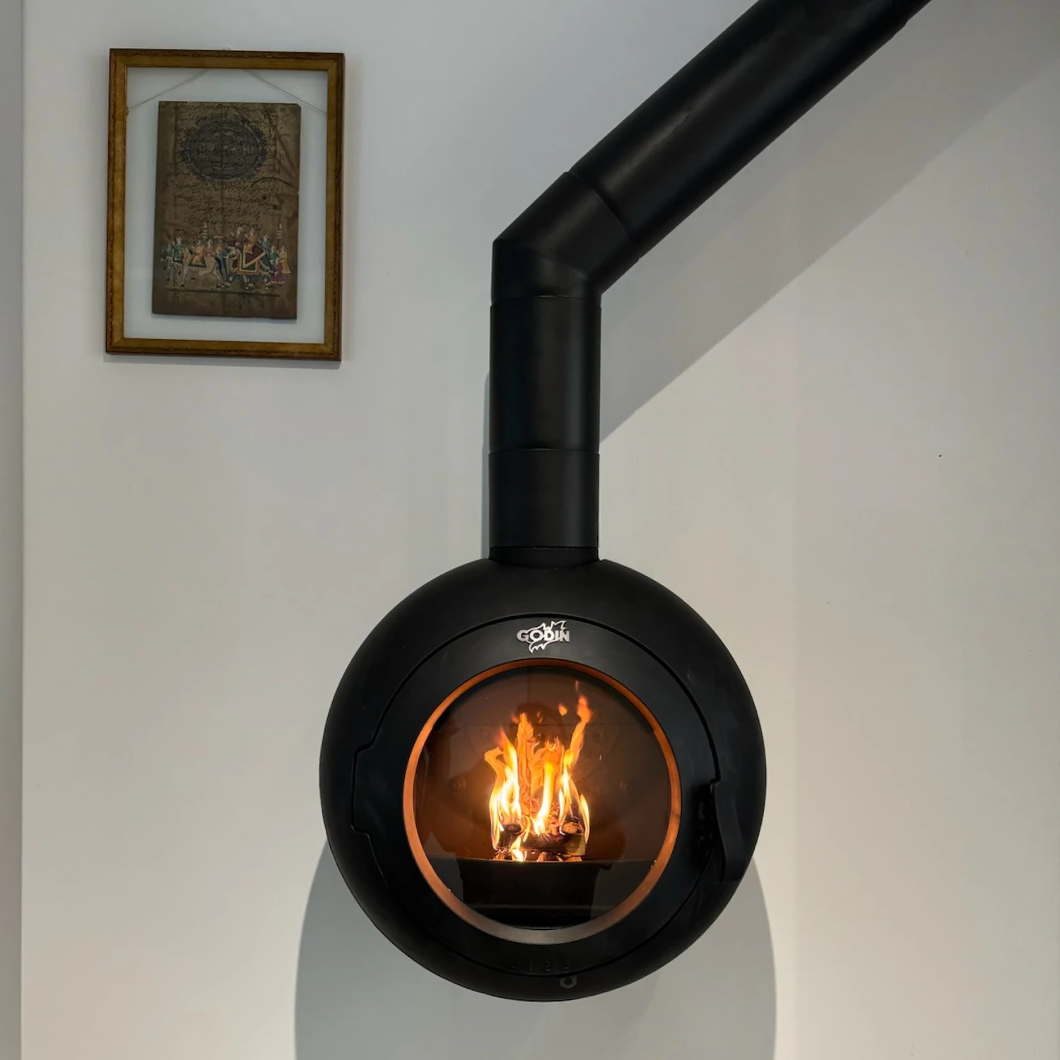 Poêle à bois rond noir avec feu visible et conduit sur un mur blanc avec un cadre au-dessus.