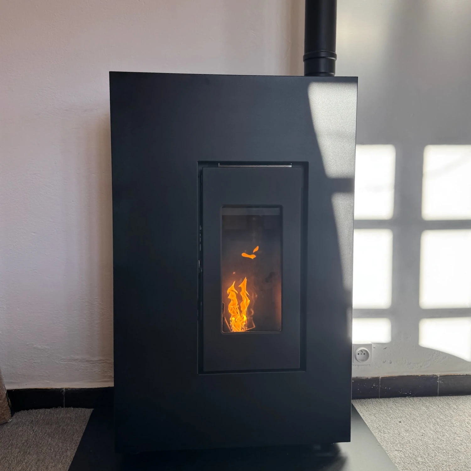 Poêle à granulés noir avec une flamme visible dans la fenêtre centrale contre un mur blanc.