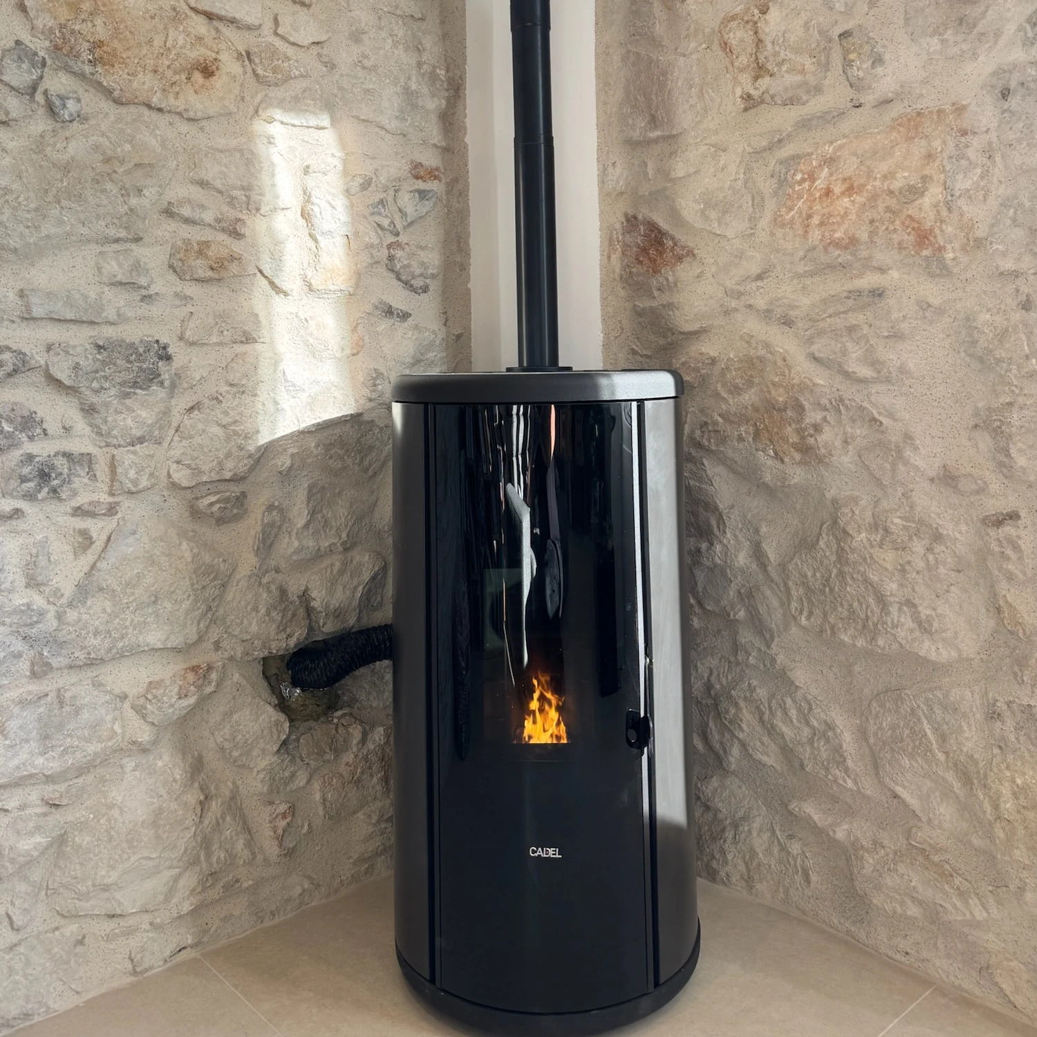 Poêle à granulés noir cylindrique en coin d’un mur en pierre avec une flamme visible.