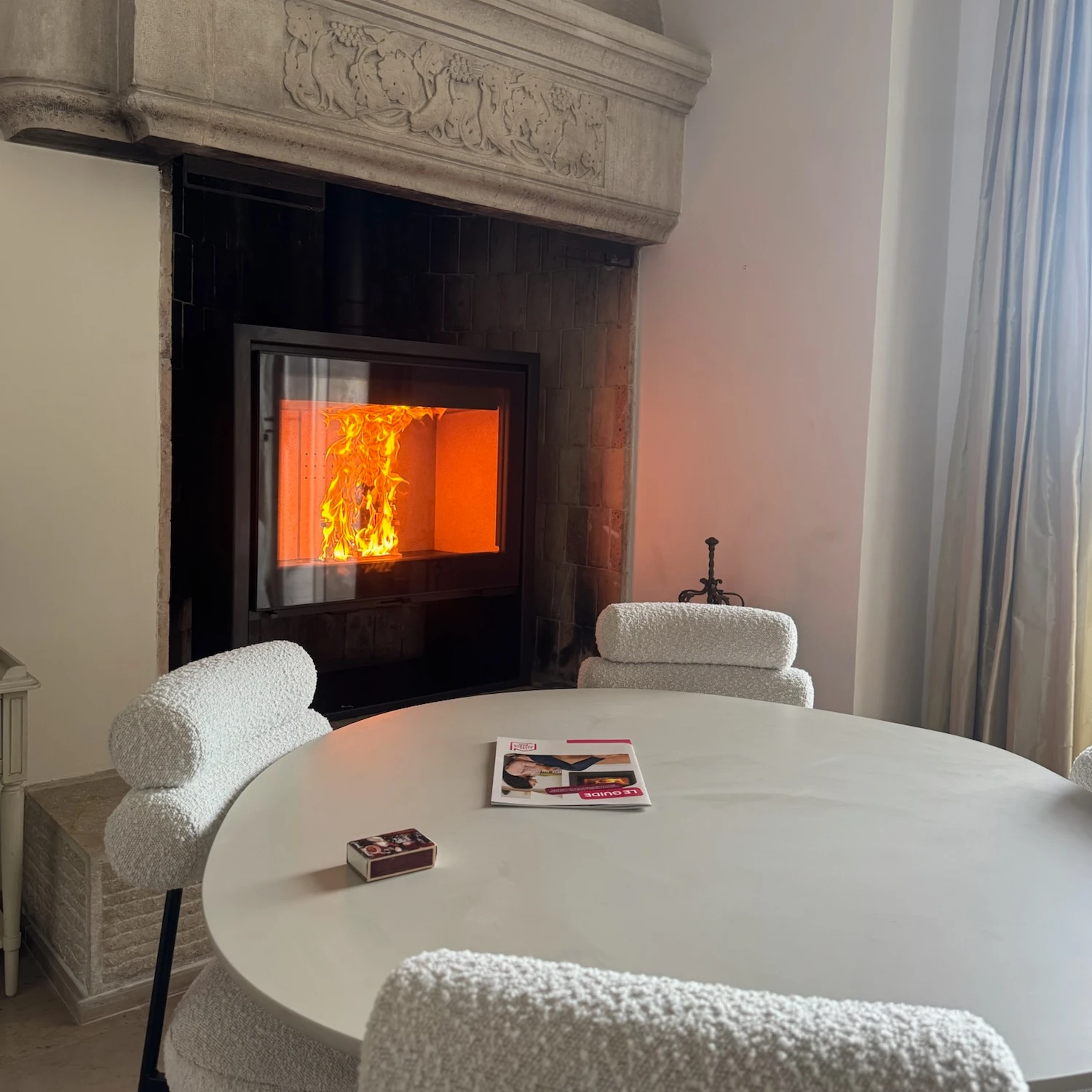 Cheminée moderne allumée dans une pièce avec table ronde et chaises blanches texturées.