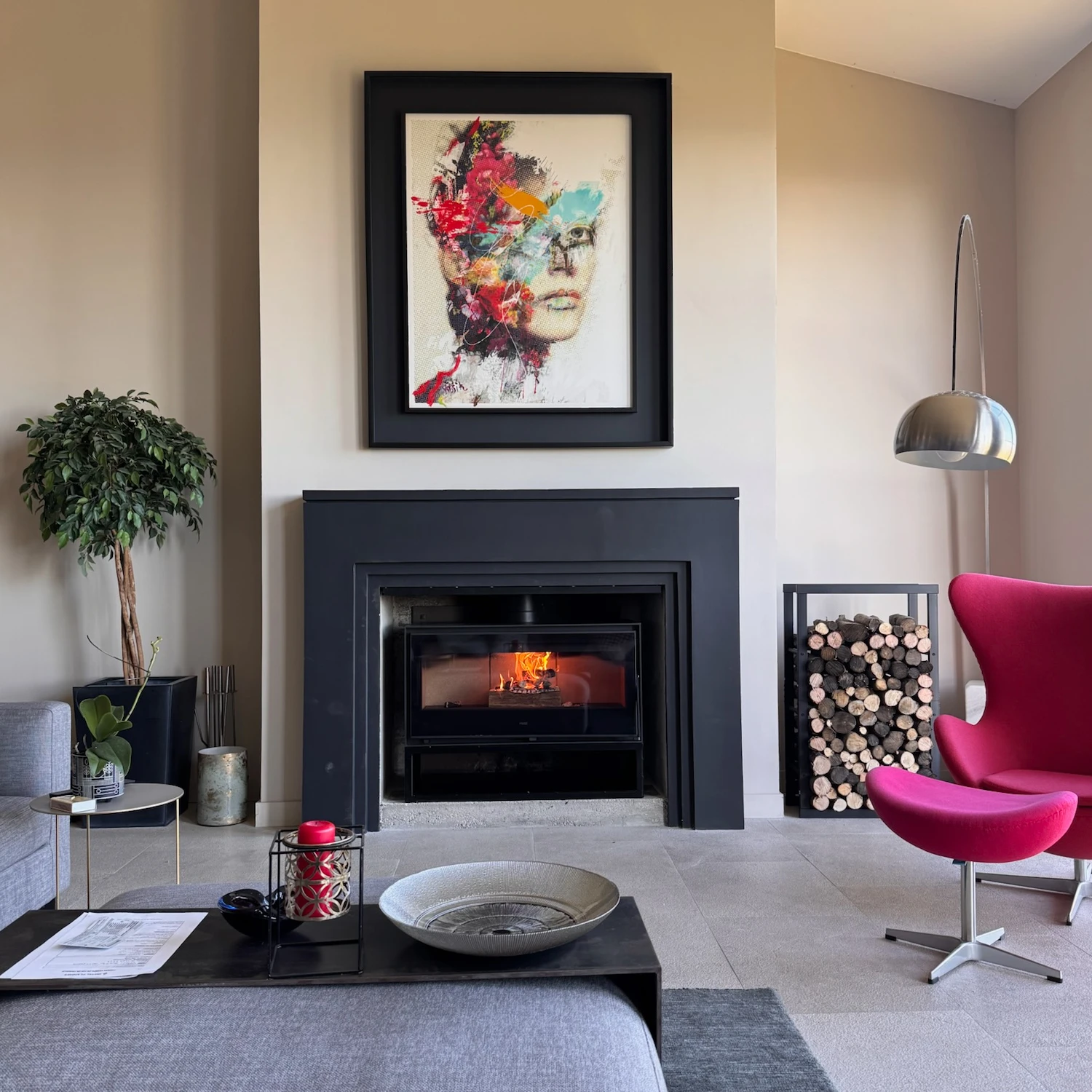 Salon moderne avec cheminée noire, fauteuil rose, table basse, tableau coloré et rangement bûches en bois.