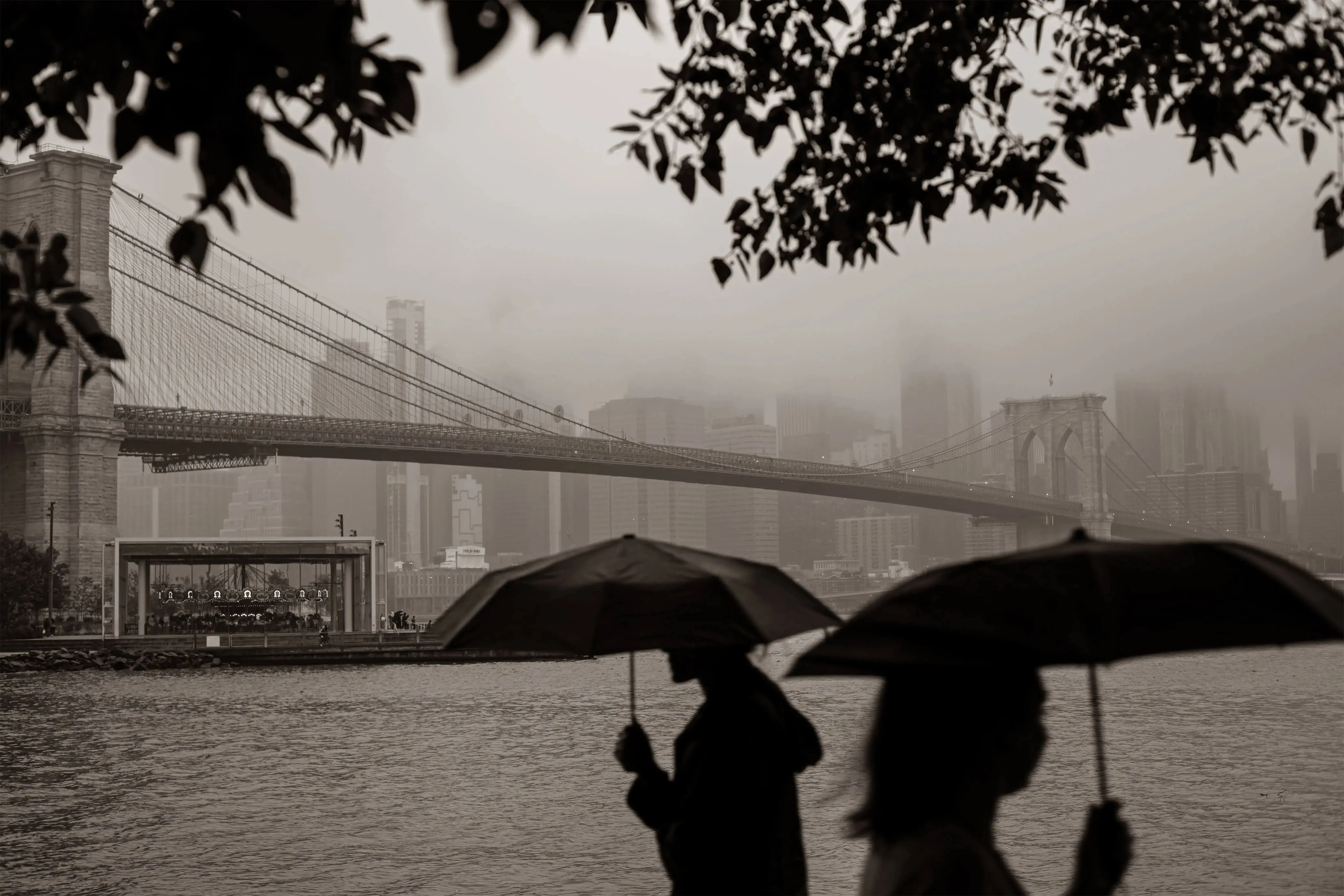A cinematic sepia-toned photograph of the Brooklyn Bridge in the rain by Daykin Dayanch, capturing the moody atmosphere of New York City through fog and silhouettes.