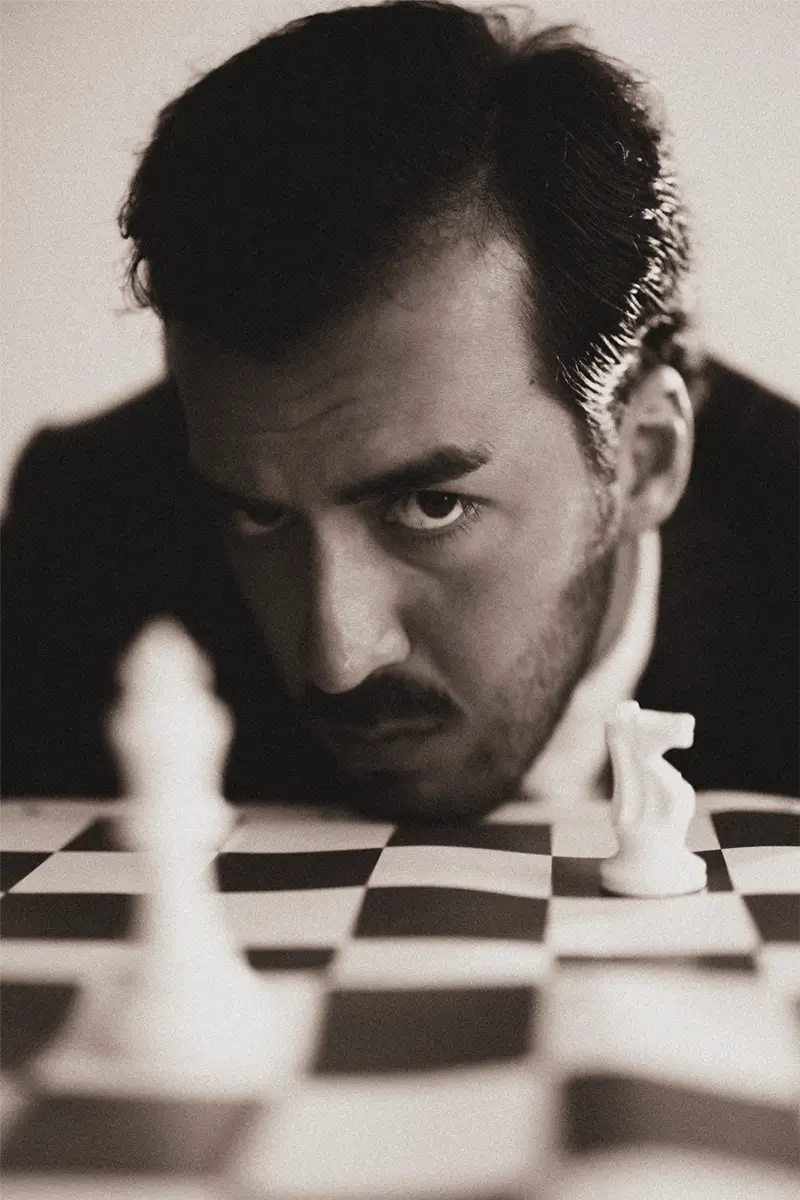 A sepia-toned self-portrait of Daykin Dayanch leaning over a chessboard. In the close-up photo, he looks down at the pieces with intense, strategic focus. The perspective is from across the board, with a white pawn and knight in the soft-focus foreground.