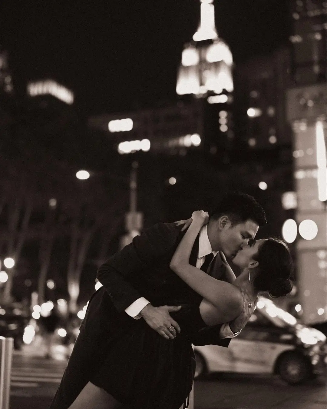 A dramatic, black-and-white photograph by Daykin Dayanch. A man in a suit dips a woman backwards for a passionate kiss on a New York City street at night. The iconic, brightly lit spire of the Empire State Building rises in the background.