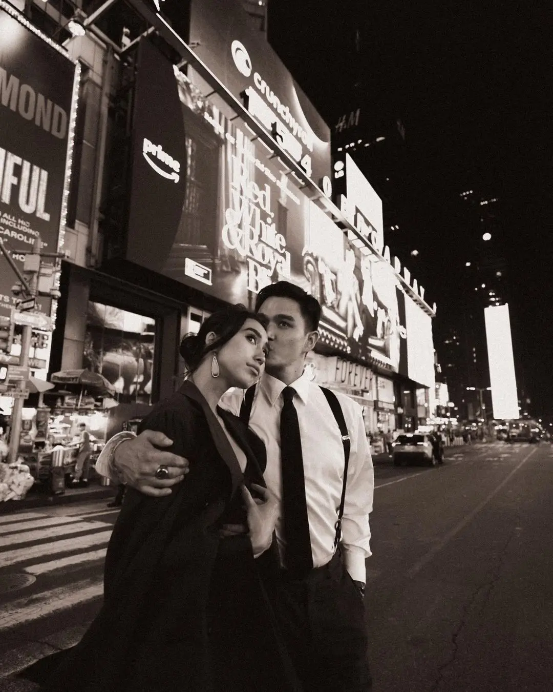 A romantic, black-and-white photograph by Daykin Dayanch. A couple stands on a crosswalk in Times Square at night. The man, wearing a shirt, tie, and suspenders, gently kisses the woman on the cheek. Behind them, the street is lit by massive digital billboards, including ads for Crunchyroll, H&M, and the movie 'Red, White & Royal Blue'.