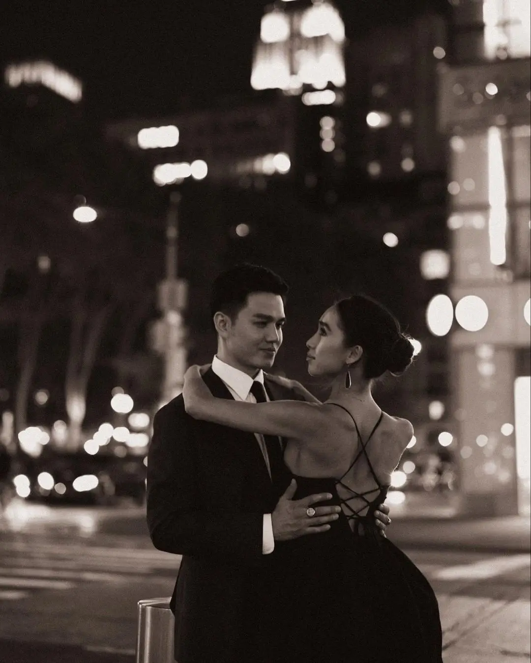 A romantic, black-and-white photograph by Daykin Dayanch. It features a formally dressed couple embracing in the middle of a city street at night. They are looking into each other's eyes, with the blurred lights of the city creating a soft, elegant background.