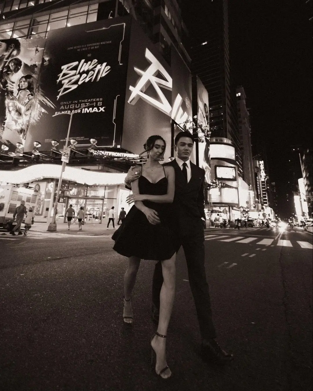 A dynamic, low-angle black-and-white photograph by Daykin Dayanch. A formally dressed couple walks across a street in Times Square at night, captured in mid-stride. Behind them, a massive digital billboard advertises the movie 'Blue Beetle,' with text showing an 'August 18' release date.