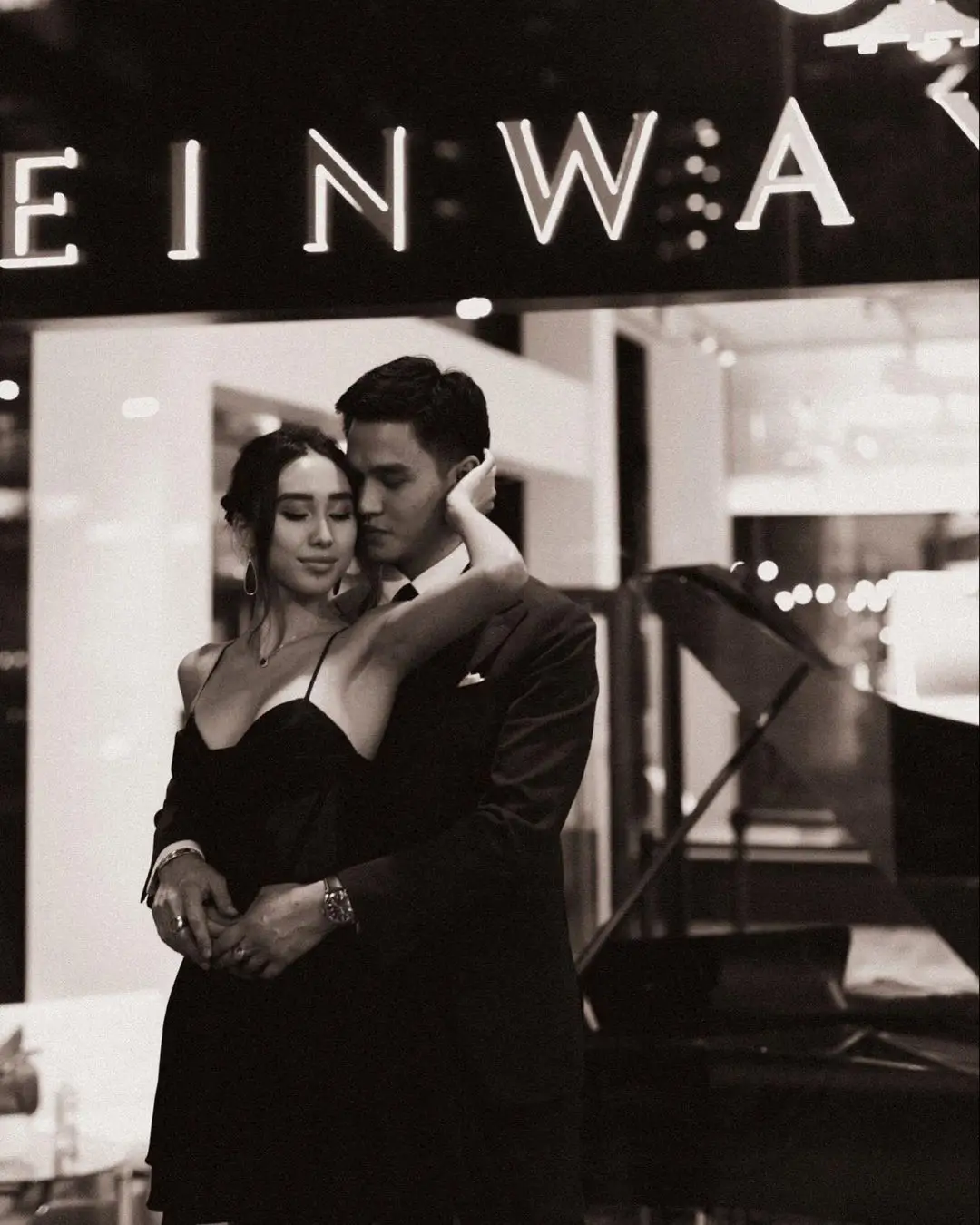 A black-and-white photograph by Daykin Dayanch, capturing a formally dressed couple in an intimate embrace. The woman, with her eyes closed, rests her arm on the man's shoulder as he holds her close. They are positioned in front of a grand piano, with part of the glowing "STEINWAY" sign visible in the background.