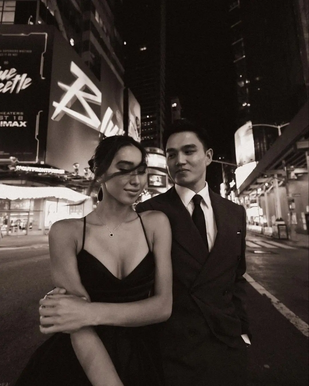 A black-and-white photograph by Daykin Dayanch. A formally dressed couple is pictured on a city street at night, surrounded by bright, commercial signs. The man, in a suit, stands behind the woman, looking at her, while she looks down with a soft smile, her arms crossed. In the background, a large digital billboard for the movie "Blue Beetle" is clearly visible.