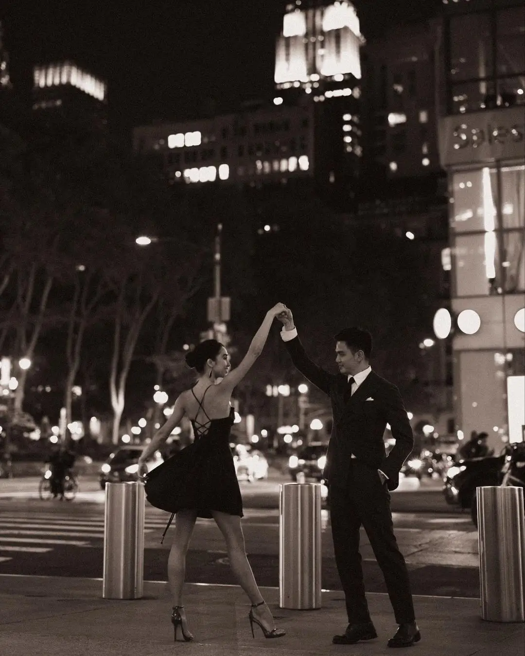 A joyous, black-and-white photograph by Daykin Dayanch. A formally dressed couple dances on a sidewalk in New York City at night. The man holds the woman's hand up as she elegantly poses on her toes in a short, backless dress. The scene is framed by street bollards, with the blurred lights of traffic and illuminated skyscrapers in the background.
