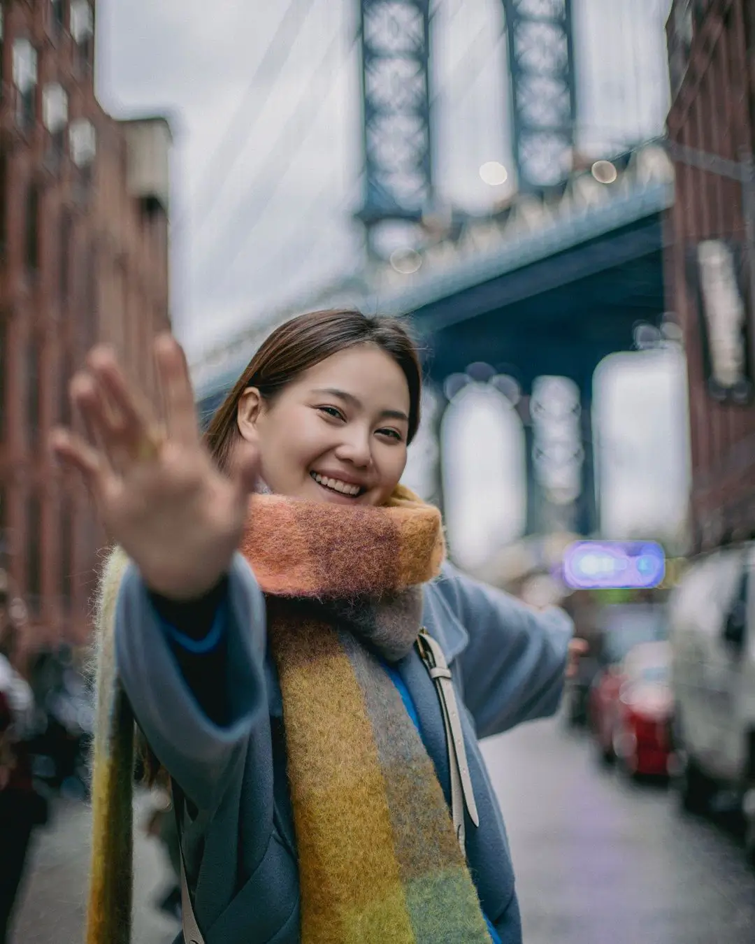 A joyful, close-up color photograph by Daykin Dayanch. A woman wearing a blue coat and a multi-colored scarf smiles broadly and waves directly at the camera. The photo is taken on the iconic DUMBO street, with the Manhattan Bridge softly blurred in the background.