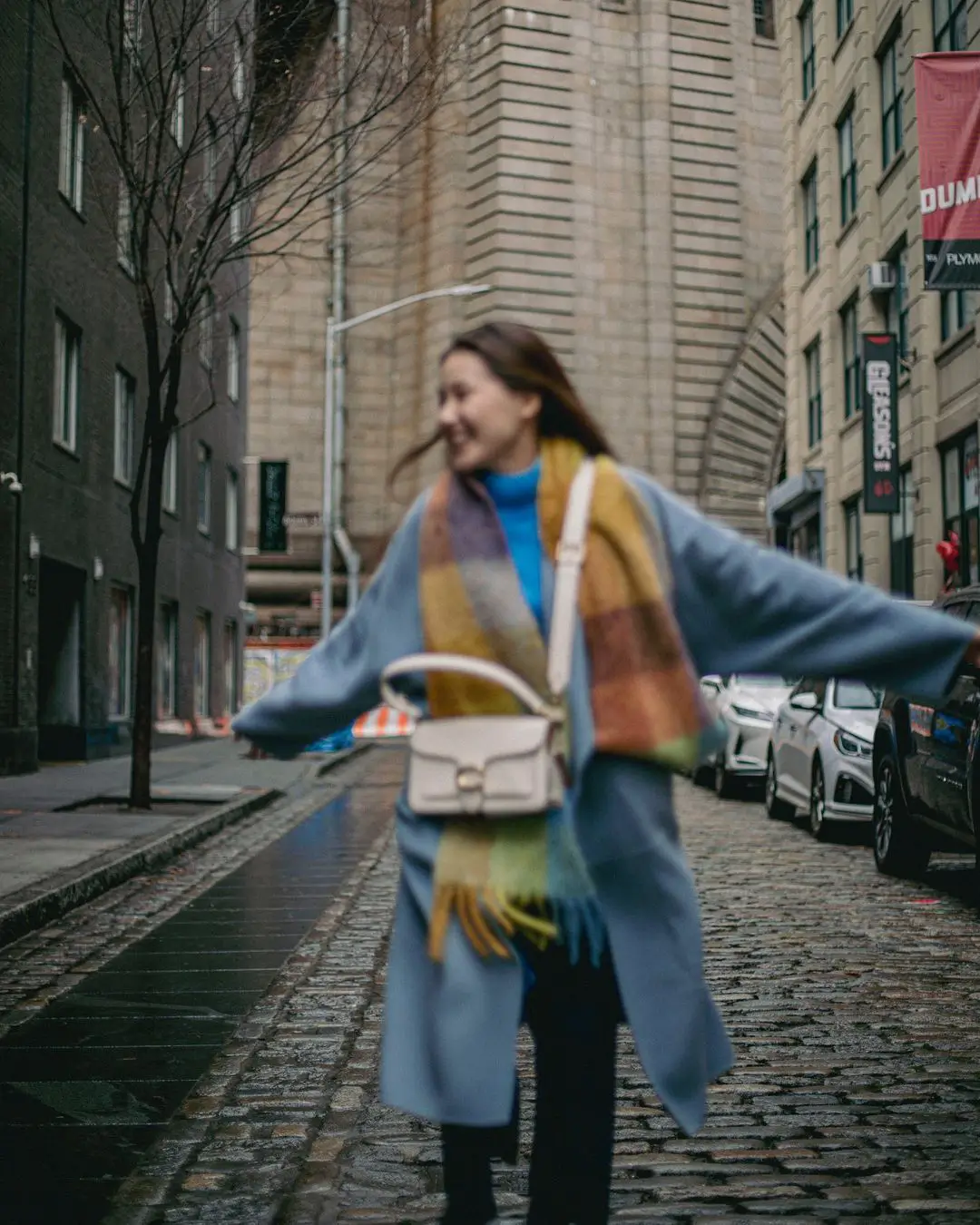 An energetic color photograph by Daykin Dayanch. The image is characterized by intentional motion blur, capturing a woman in a blue coat and colorful scarf as she spins with arms outstretched on a cobblestone street in DUMBO. Her joyful movement creates the blur, and a sign for "Gleason's" is visible in the background.