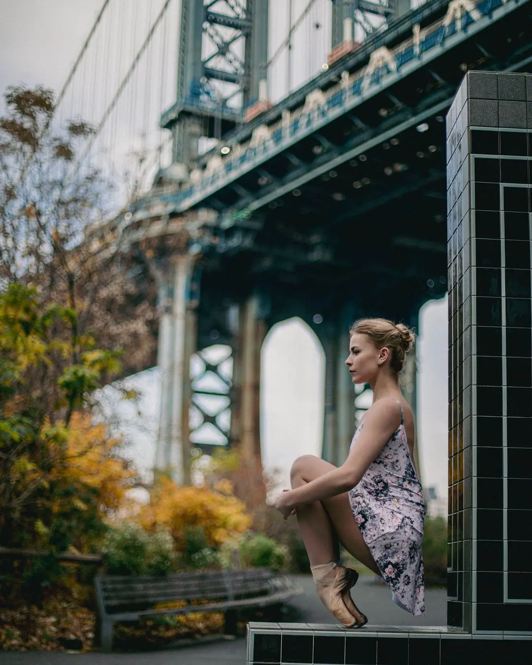 A color photograph by Daykin Dayanch. A ballet dancer in a patterned leotard and pointe shoes is crouched in profile on a tiled pillar in a DUMBO park. She gazes forward with a calm expression. The massive steel and stone structure of the Manhattan Bridge and trees with autumn foliage compose the background.