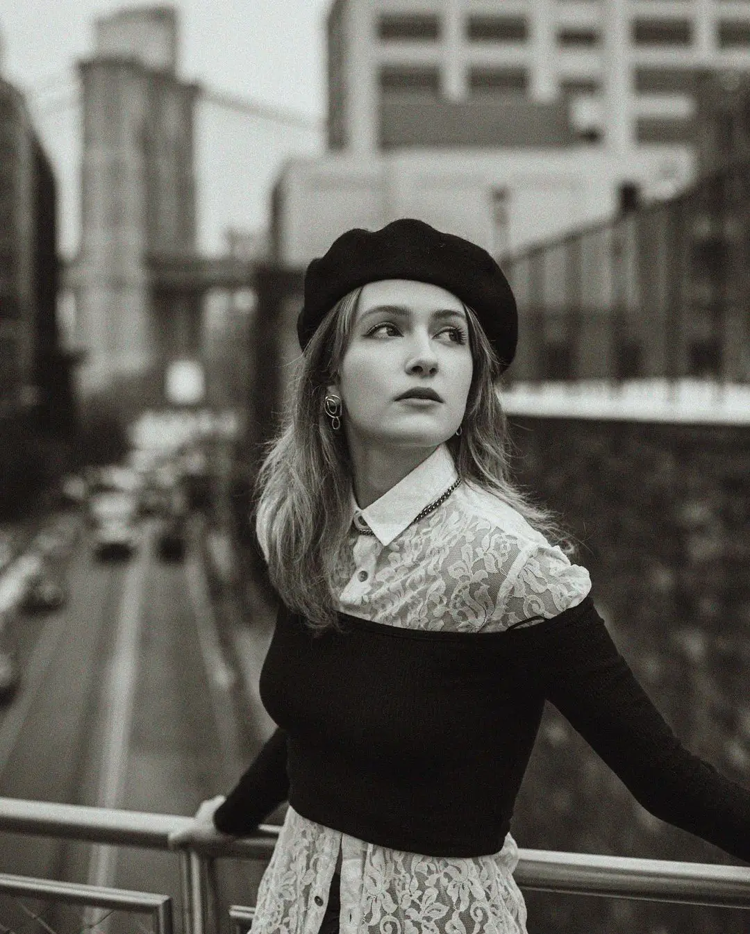 A black-and-white photograph by Daykin Dayanch. A young woman wearing a black beret and a lace shirt stands on an overpass of Squibb Park Bridge, looking off to the side. In the background, the iconic cables and stone towers of the Brooklyn Bridge are visible, along with traffic on the road below.